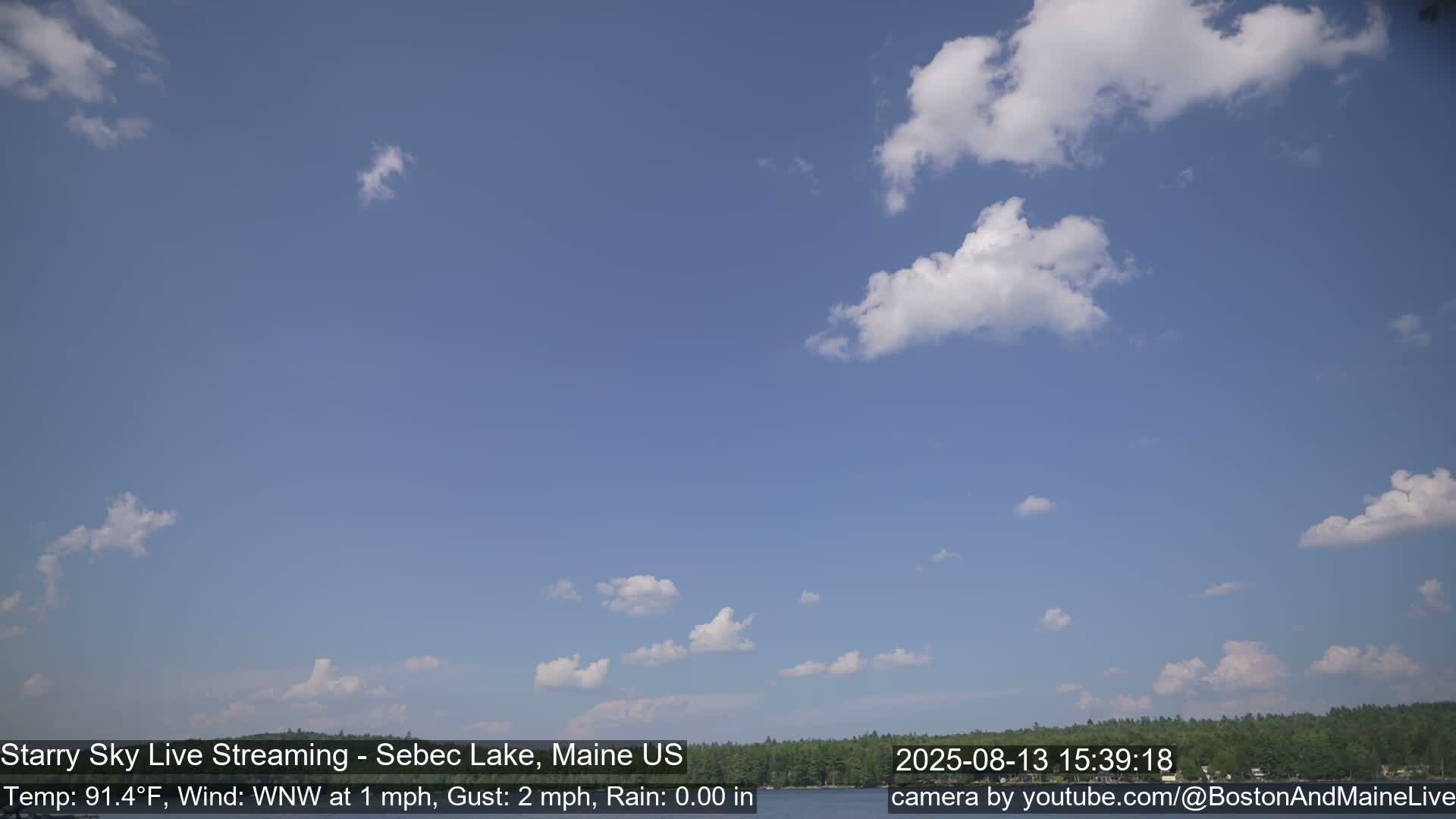 A mostly clear blue sky with scattered cumulus clouds is visible over a calm lake and tree line.