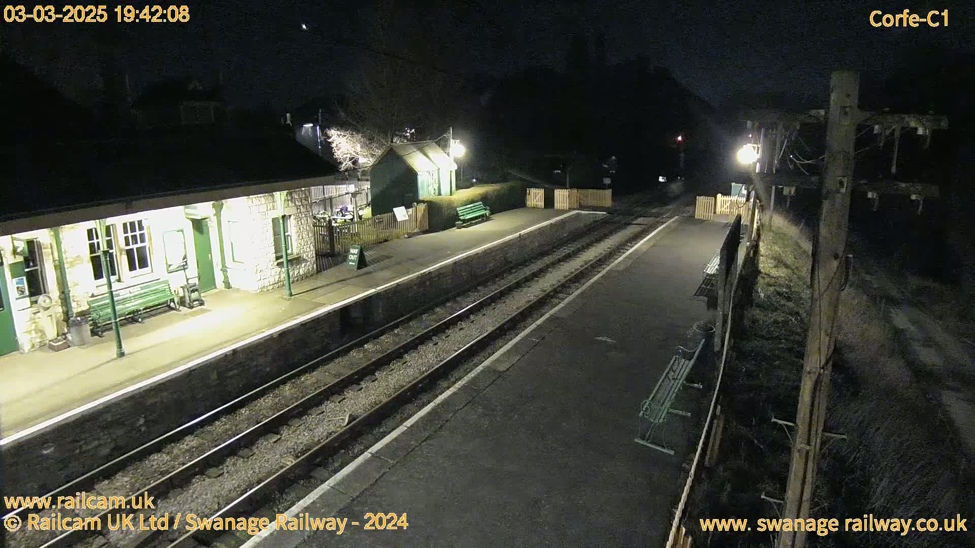 A deserted train station at night, illuminated by artificial lights, with train tracks extending into the darkness.