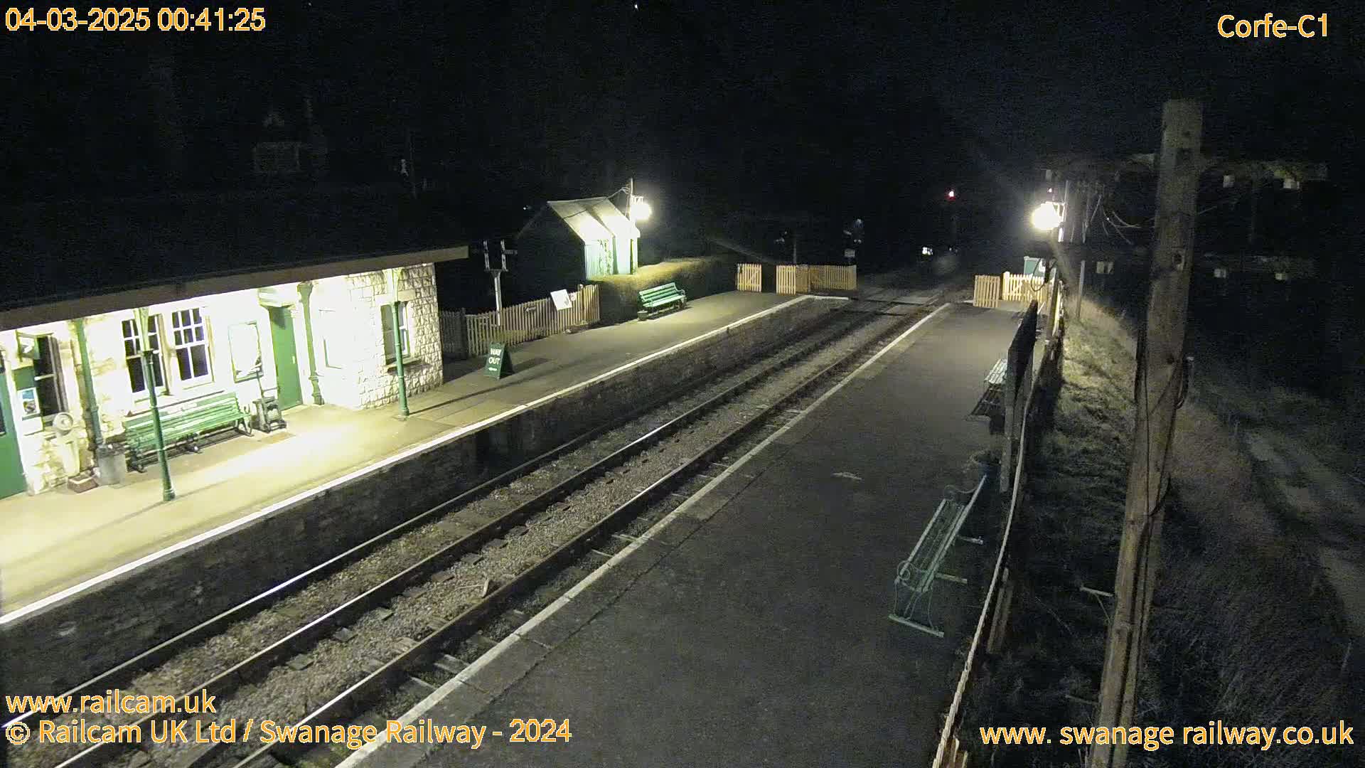 A nighttime view of a deserted train station platform with two sets of tracks, illuminated by several lights.
