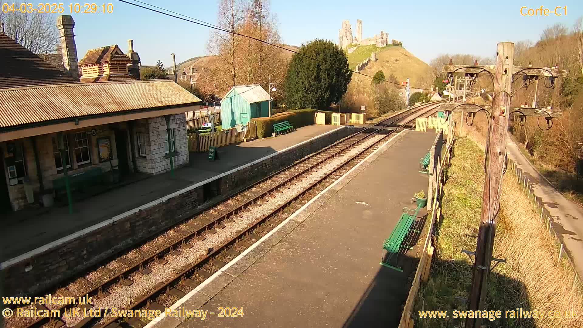 A train station platform with a stone building and a view of a castle ruin on a sunny hill in the background.