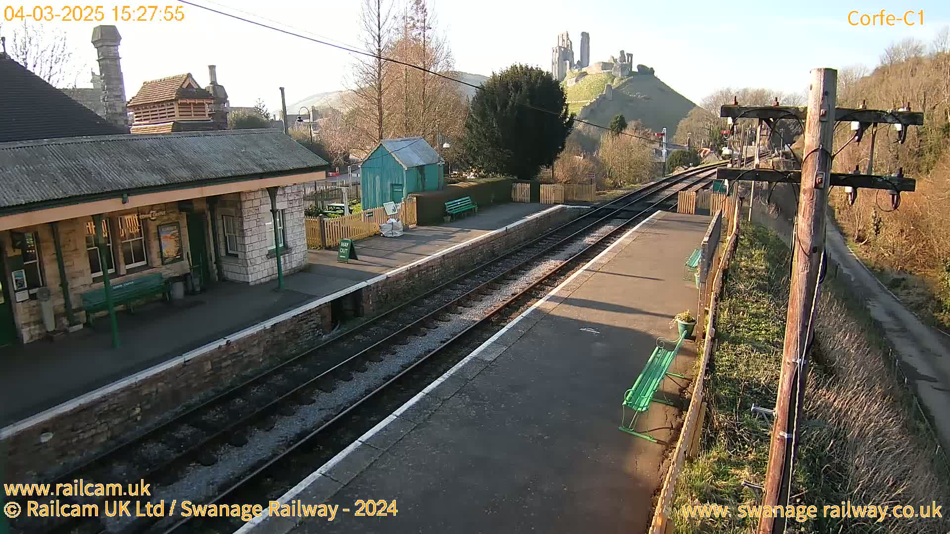 A train station platform with two train tracks is shown, a stone building is to the left and a hill with a castle ruin is visible in the background under a sunny sky.