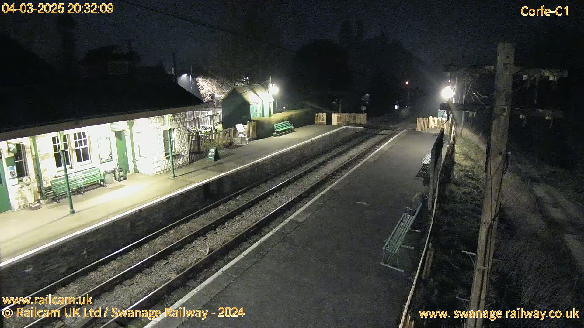 A deserted train station platform at night, illuminated by artificial lights, with train tracks extending into the darkness.