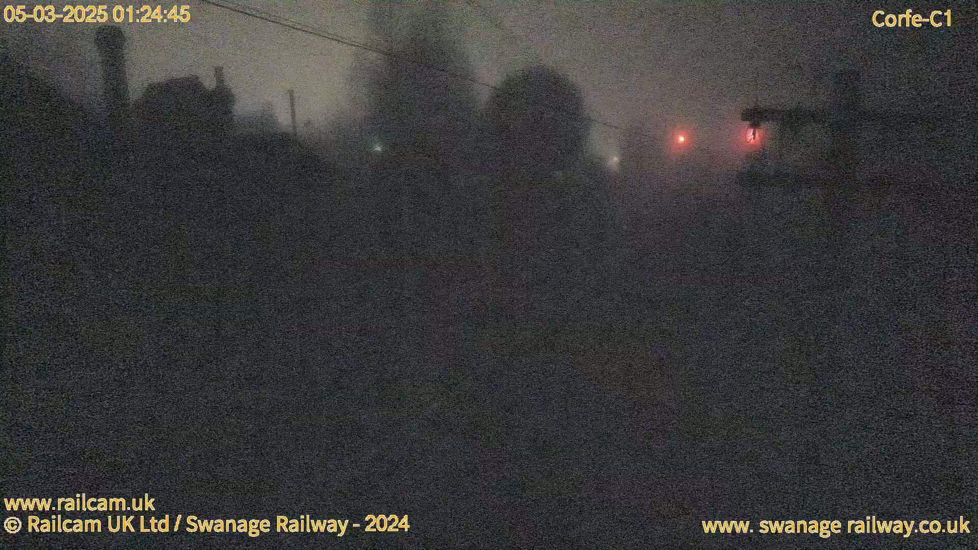A dark, foggy nighttime scene shows indistinct shapes of buildings and objects in the distance, with a few faint red lights visible.