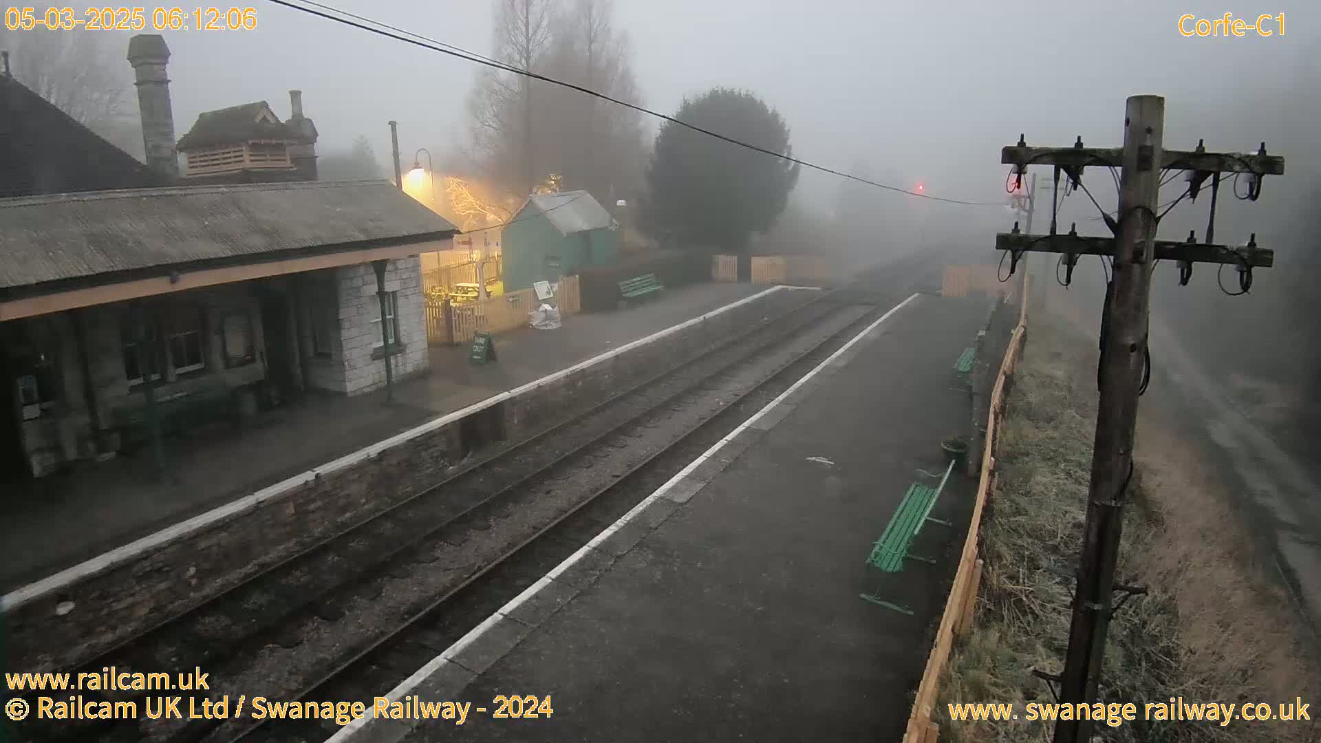 A foggy view of a train station platform with two sets of train tracks, a stone building, and several benches.