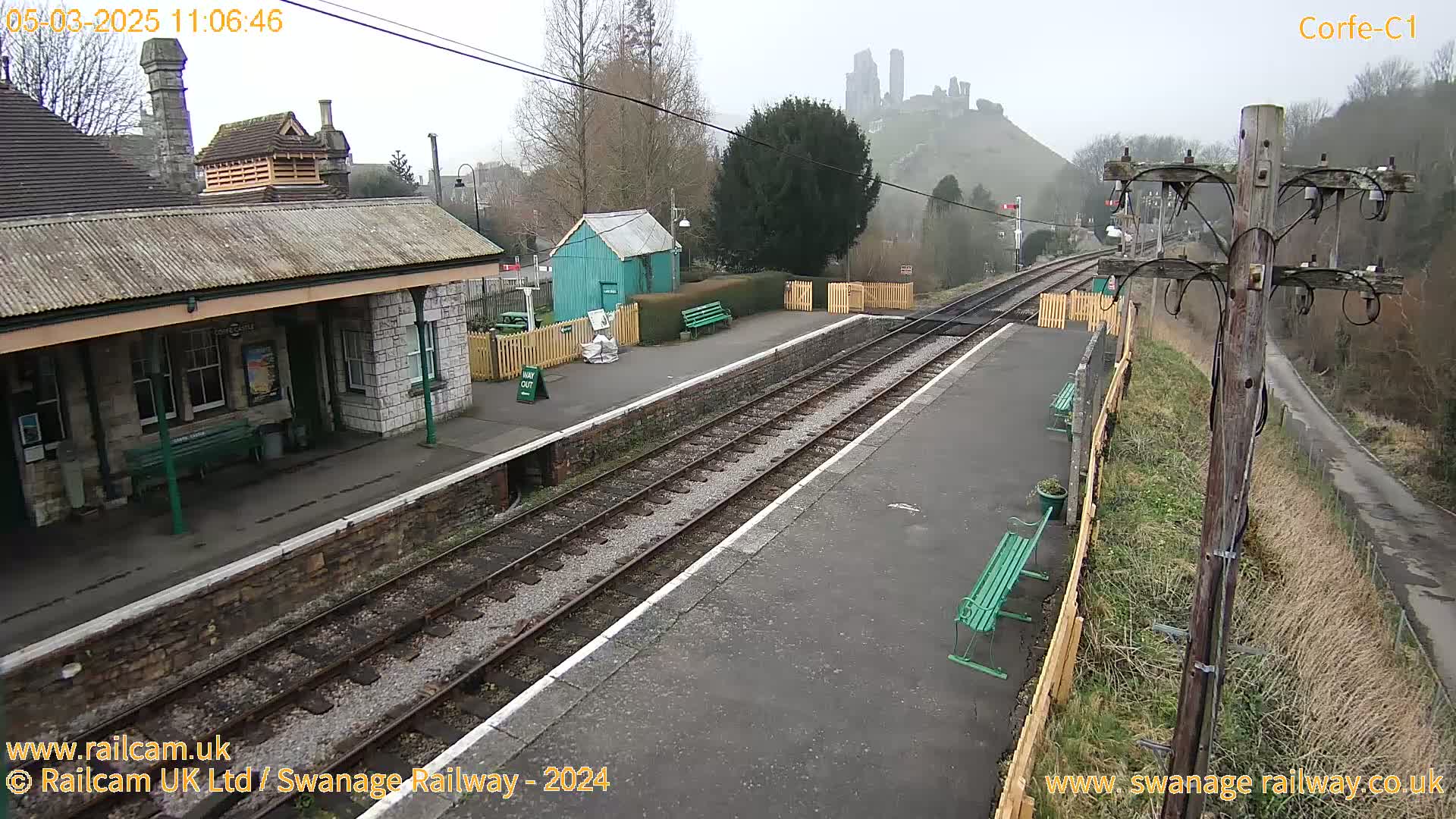 A train station platform with a stone building, a small teal shed, and benches overlooks a hill with a castle ruin under an overcast sky.