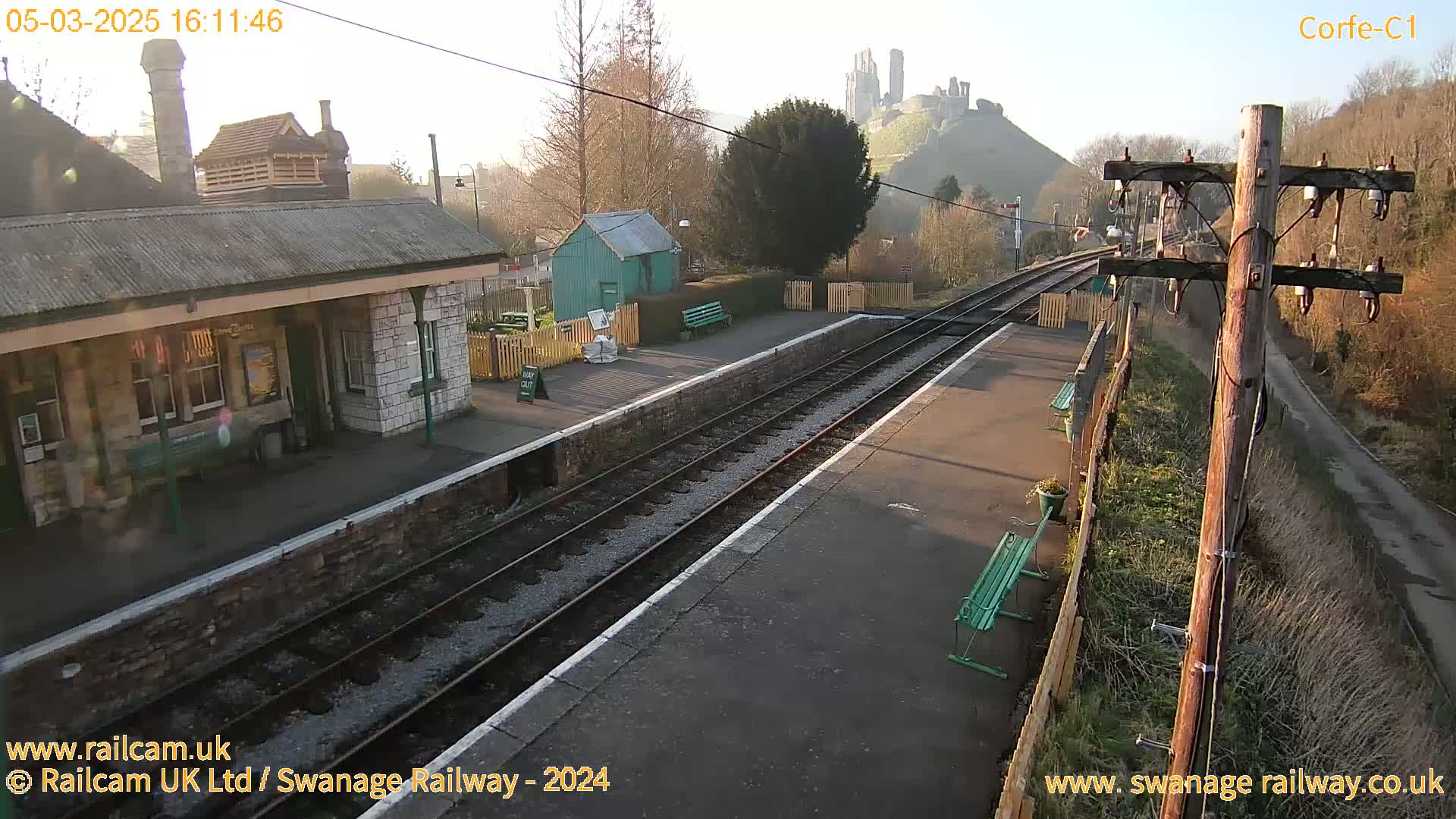 A train station platform with a stone building and a castle visible in the distance on a sunny day.