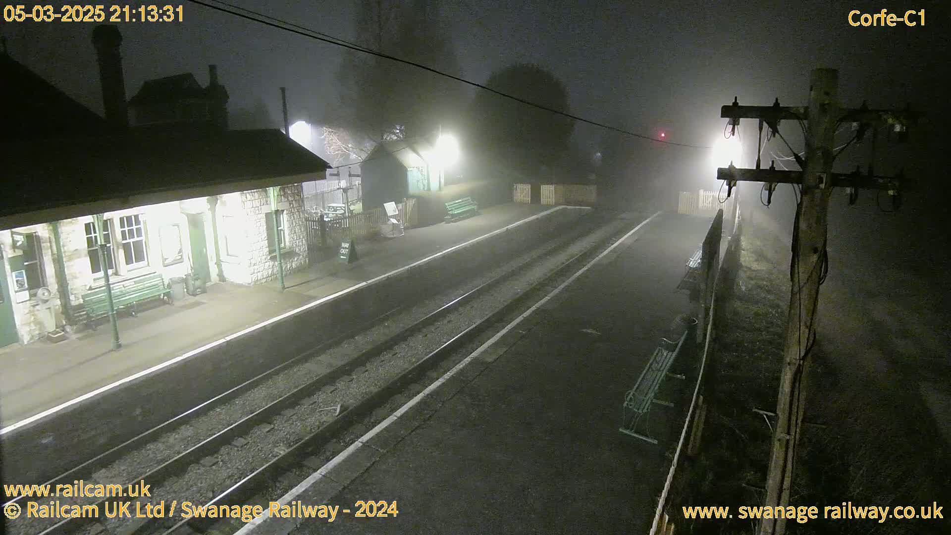 A fog-shrouded train station at night, with a building and platform visible under dim lighting.
