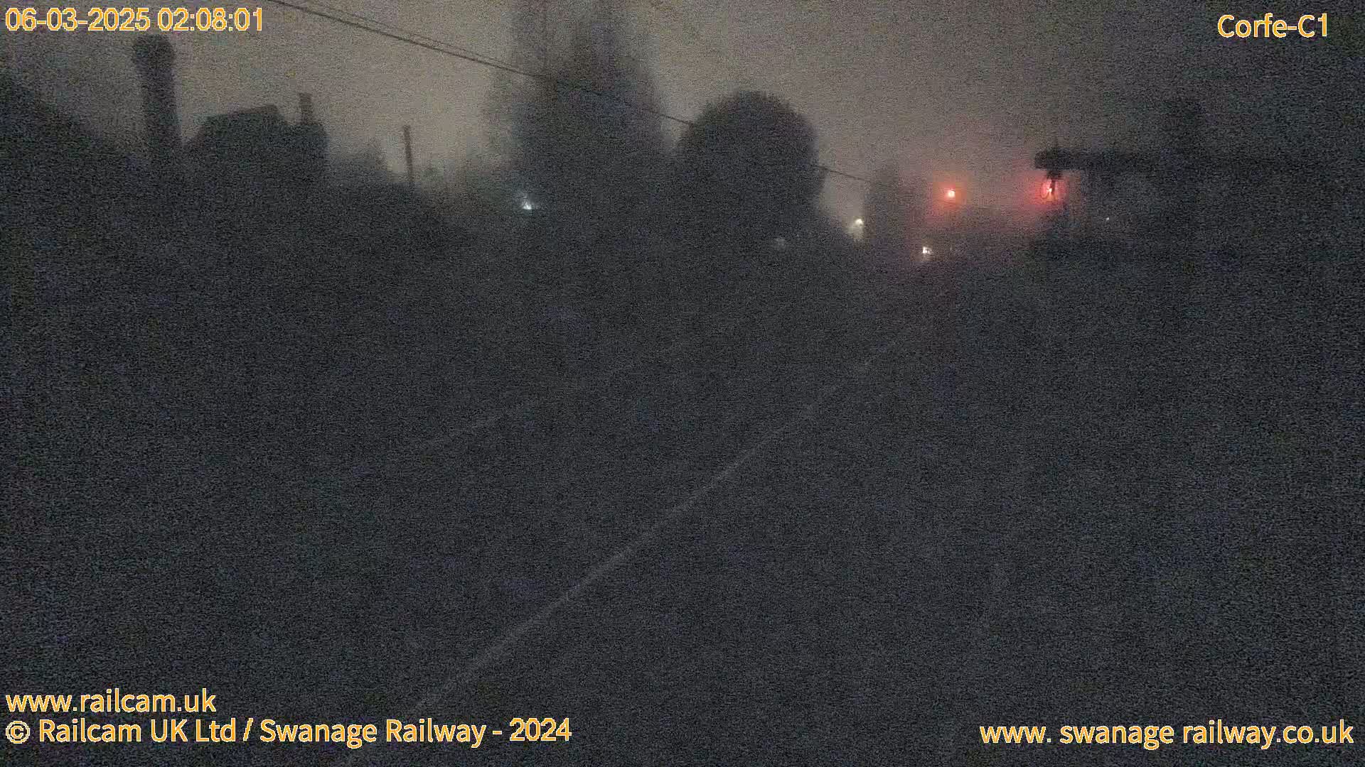 A dark, foggy nighttime scene shows a railway track with dimly lit structures and vegetation in the background.