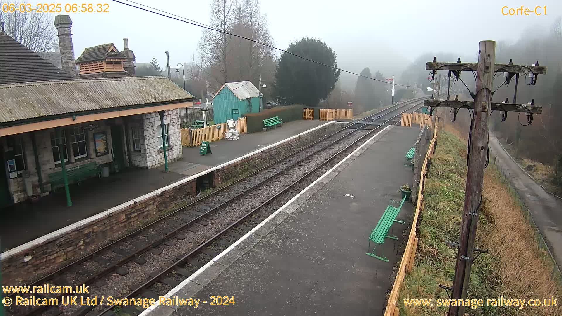 A foggy view of a train station platform with a stone building, a small green shed, and benches.