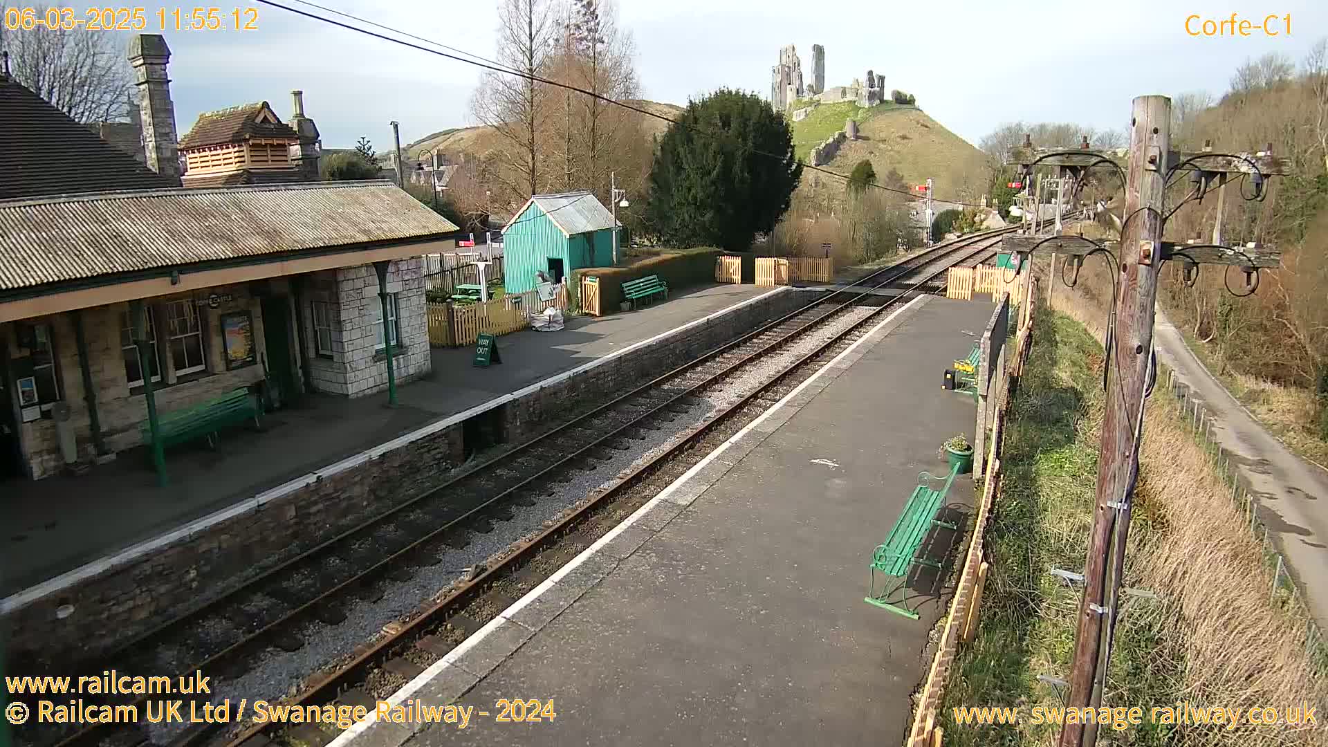 A sunny view of a train station platform with a stone building, a small green shed, and a hill with castle ruins in the background.