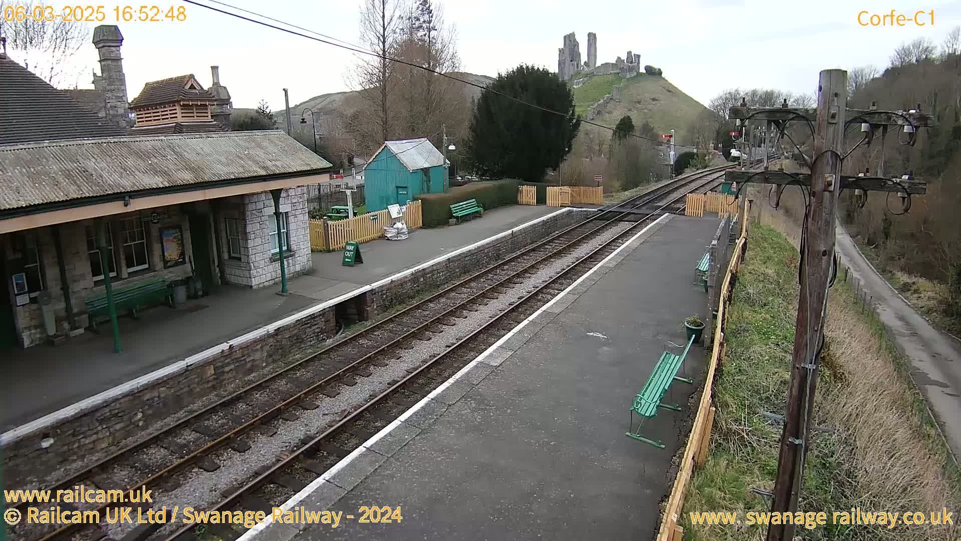 A train station platform with two sets of tracks, a stone building, and a small green building, all under a mostly clear sky with a hill and castle ruins visible in the background.