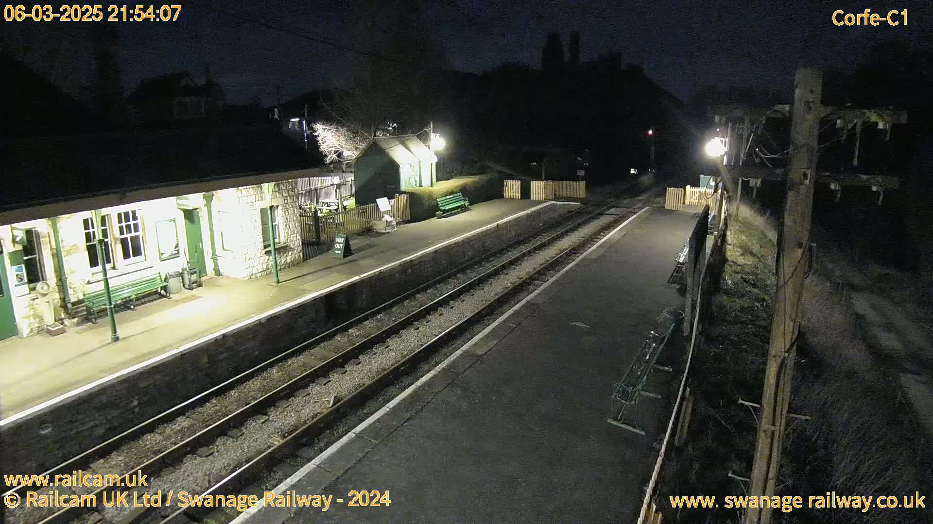 A deserted train station at night, illuminated by artificial lights, with two sets of train tracks extending into the darkness.