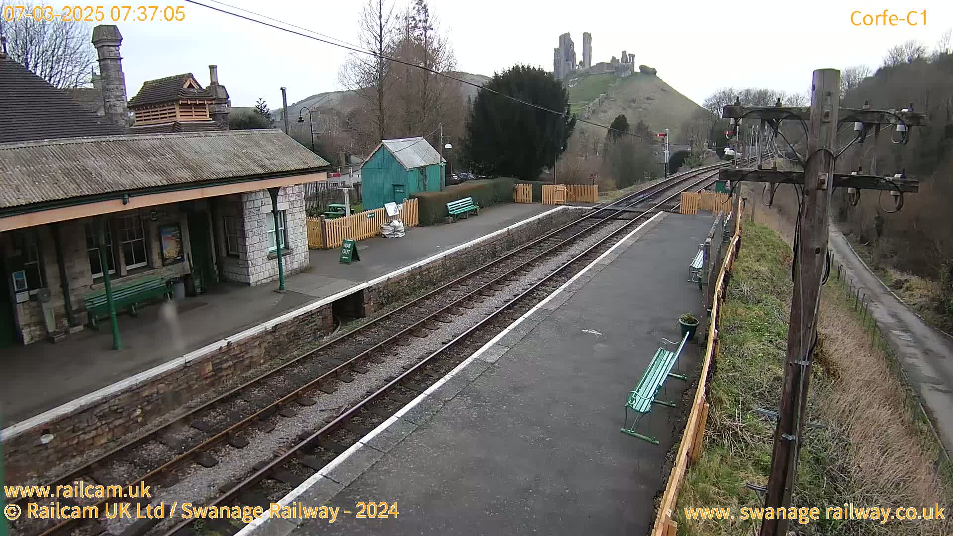 A train station platform with a stone building and a distant hilltop castle is visible under an overcast sky.