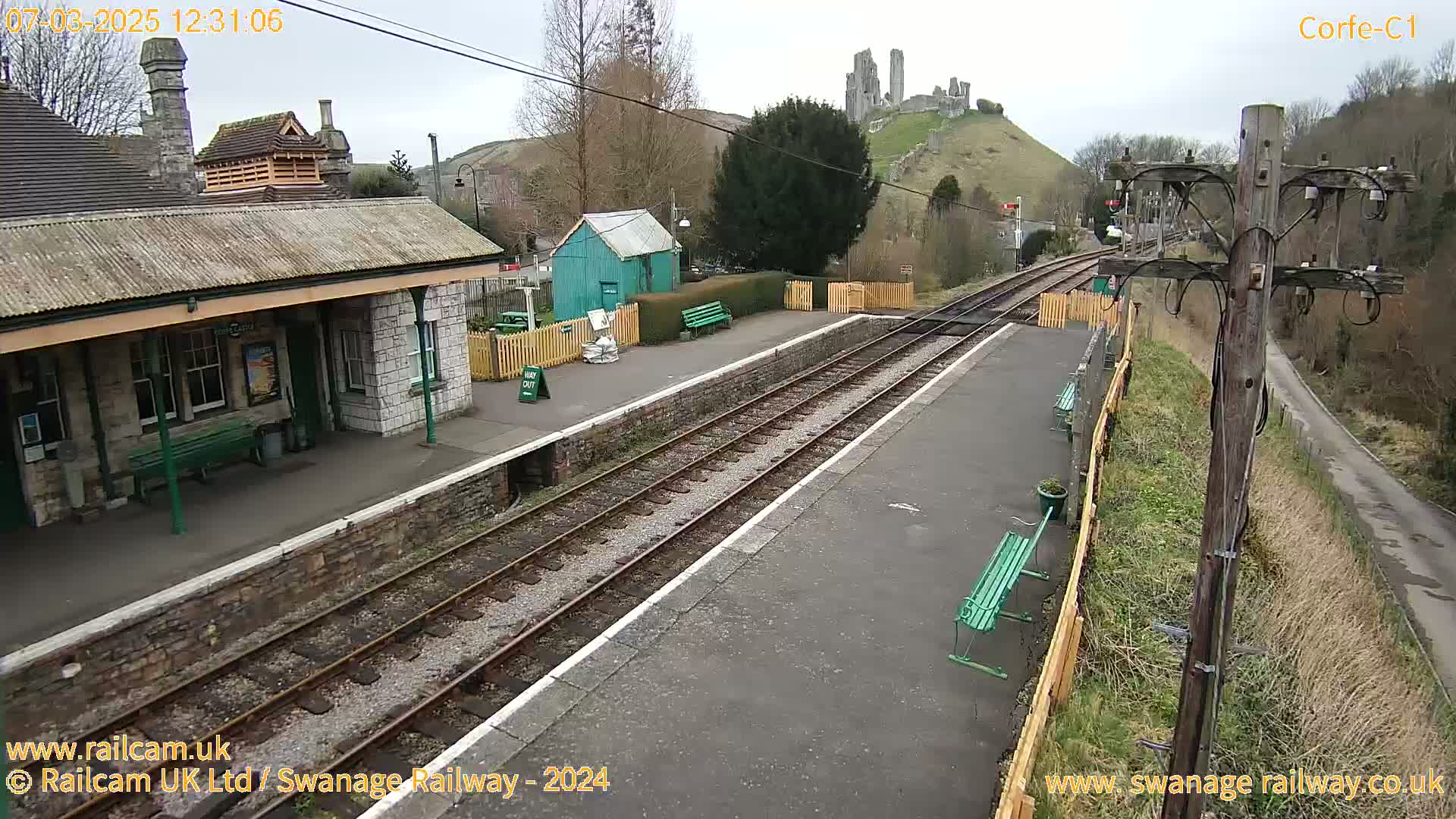 A train station with a stone building and a small green shed, situated on a railway line with a hill and castle ruins in the background on an overcast day.
