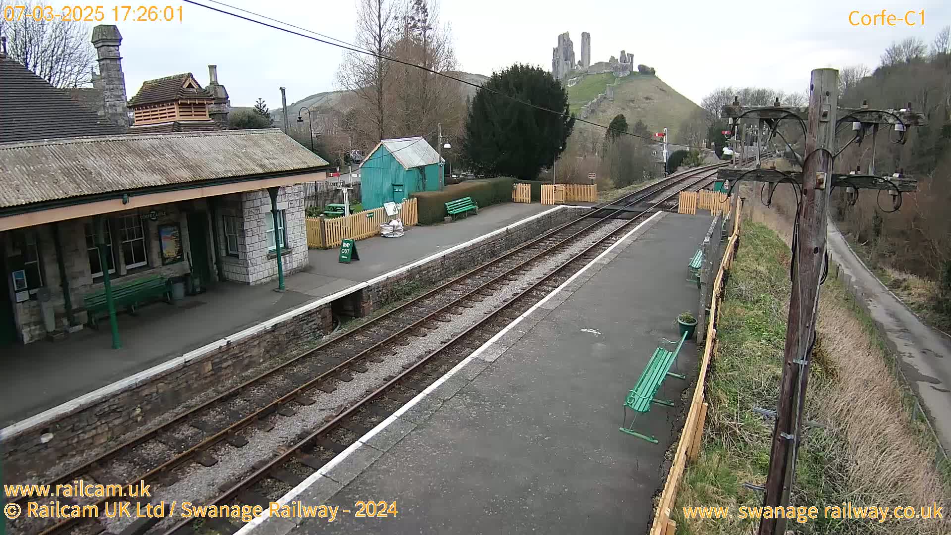 A train station with a stone building and a green shed, overlooking a hill with a castle ruin under an overcast sky.