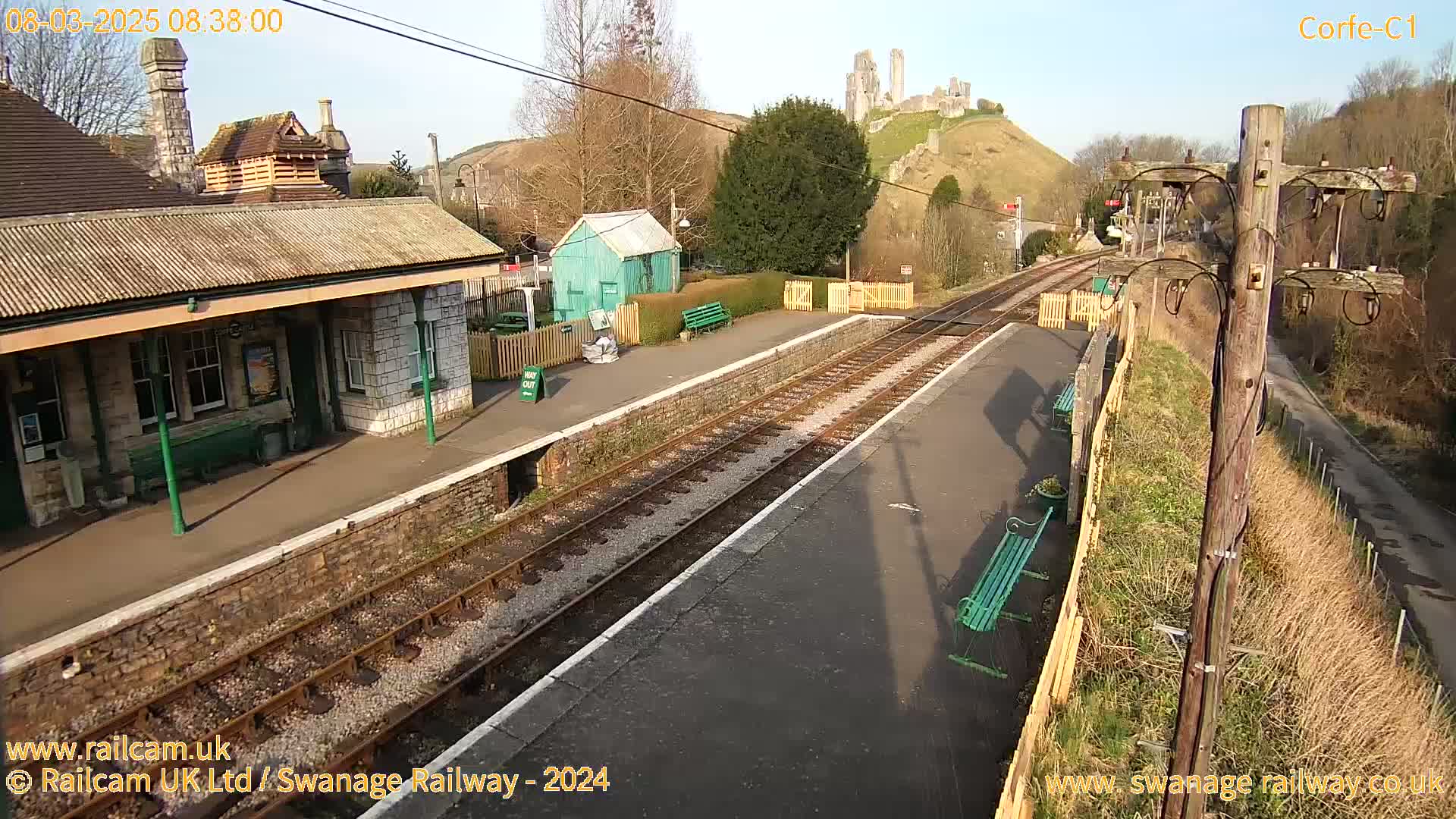 A train station platform with a stone building and a distant hilltop castle under sunny skies.