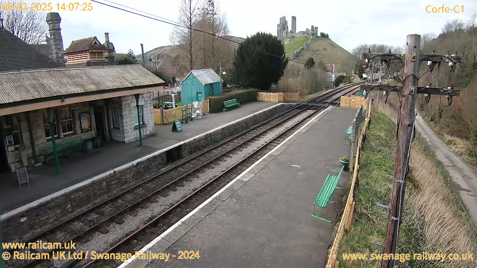 A train station with a stone building, a small green shed, and train tracks, overlooks a hill with a castle ruin under a partly cloudy sky.