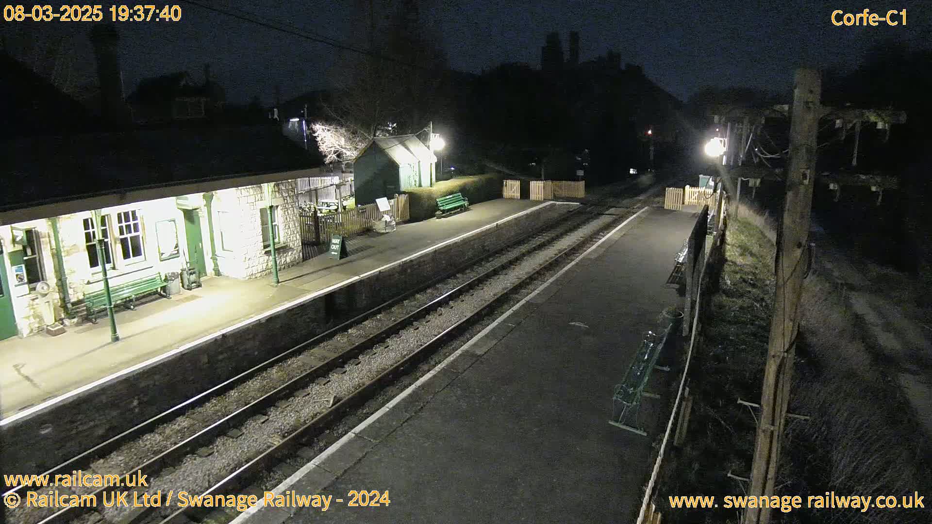 A nighttime view of a deserted train station platform with two sets of tracks, illuminated by several lights, under a dark sky.