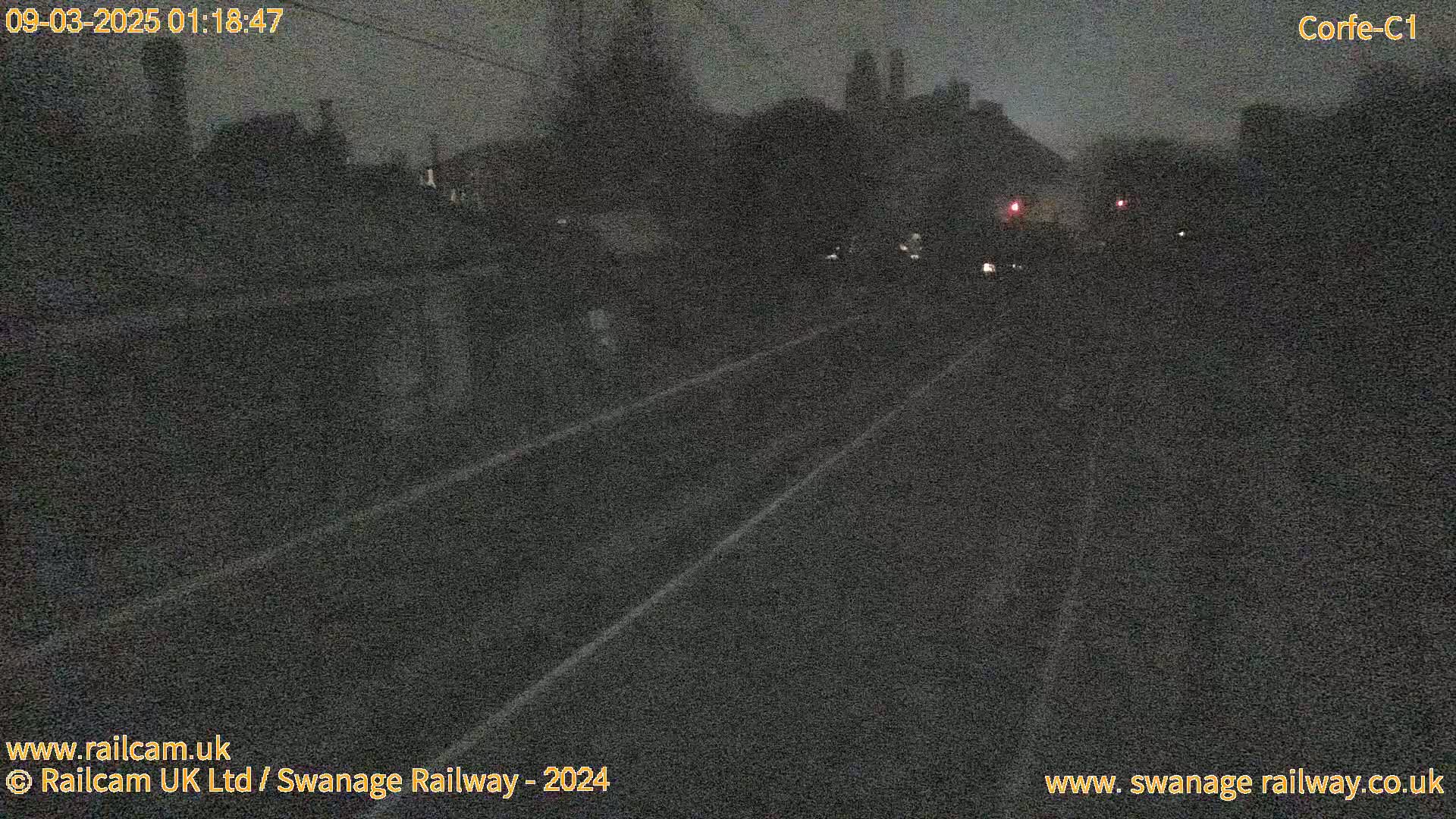 A dark, nighttime view of a railway track, with indistinct structures and faint lights in the background.