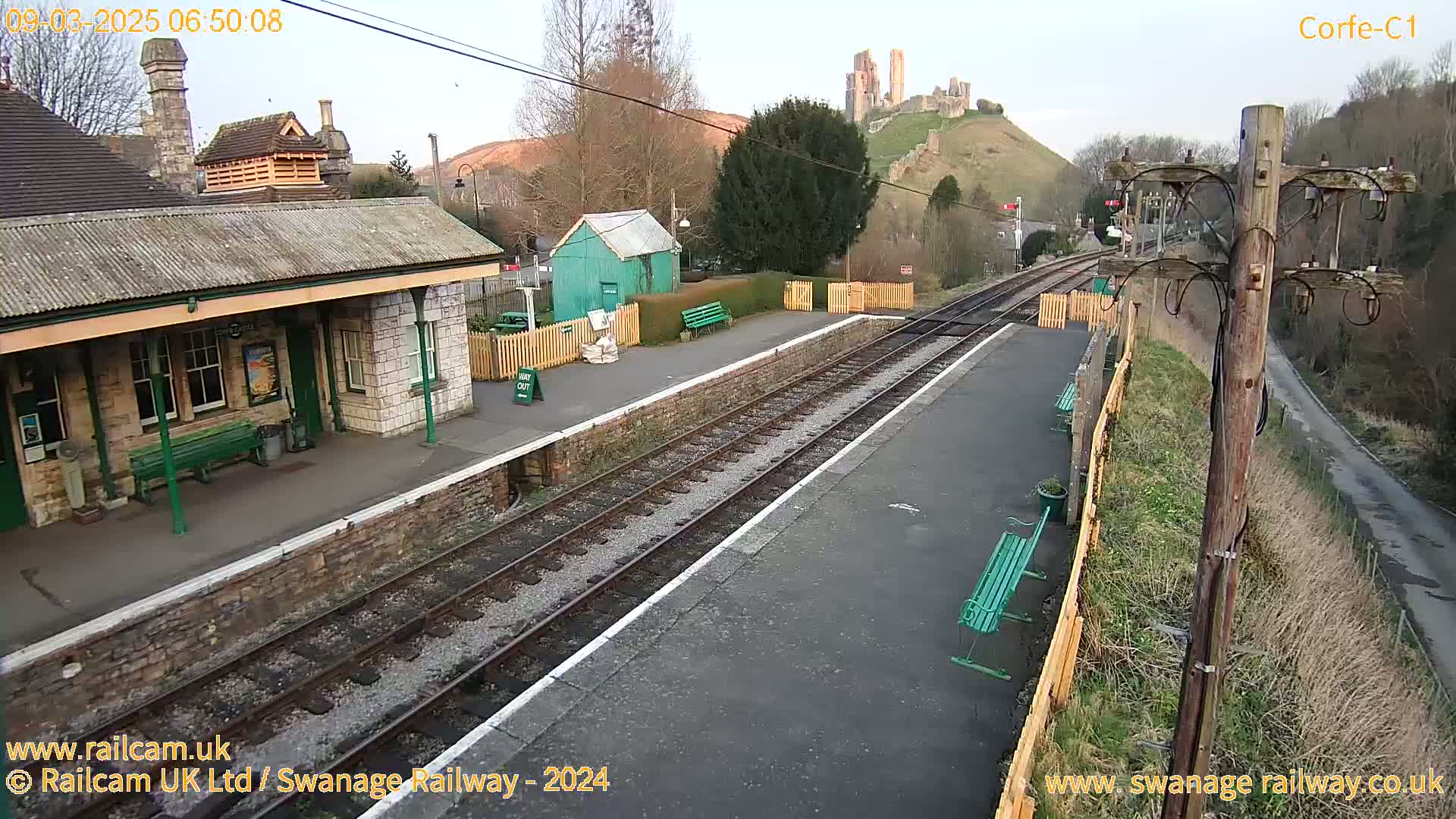 A train station platform with a stone building and green benches, situated on a hill overlooking a castle ruin under a partly cloudy sky.