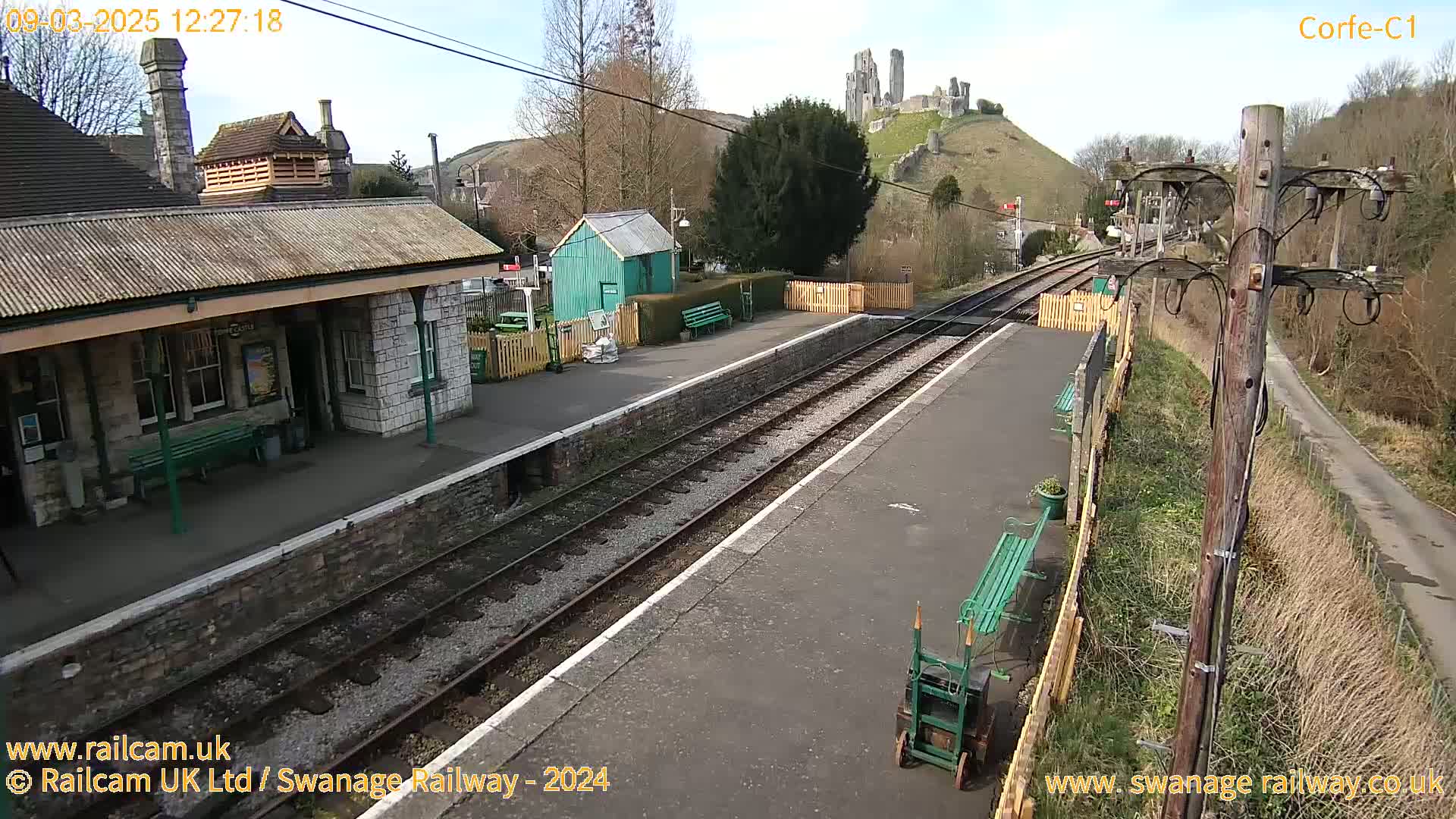A train station platform with train tracks, a stone building, and a small green shed, all under a sunny sky, with a castle ruin visible on a hill in the background.