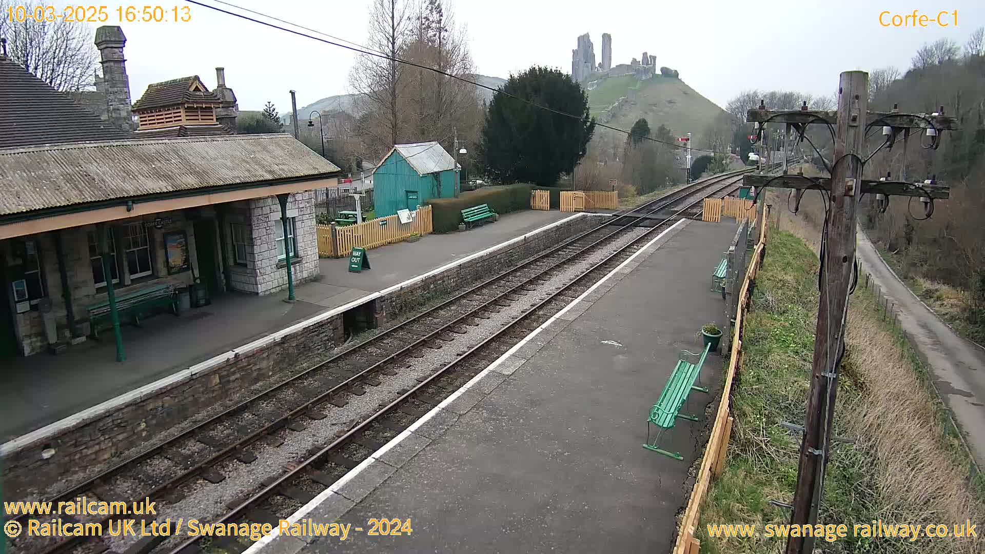 A train station with a stone building and benches, railroad tracks, and a hill with a castle ruin in the background under an overcast sky.