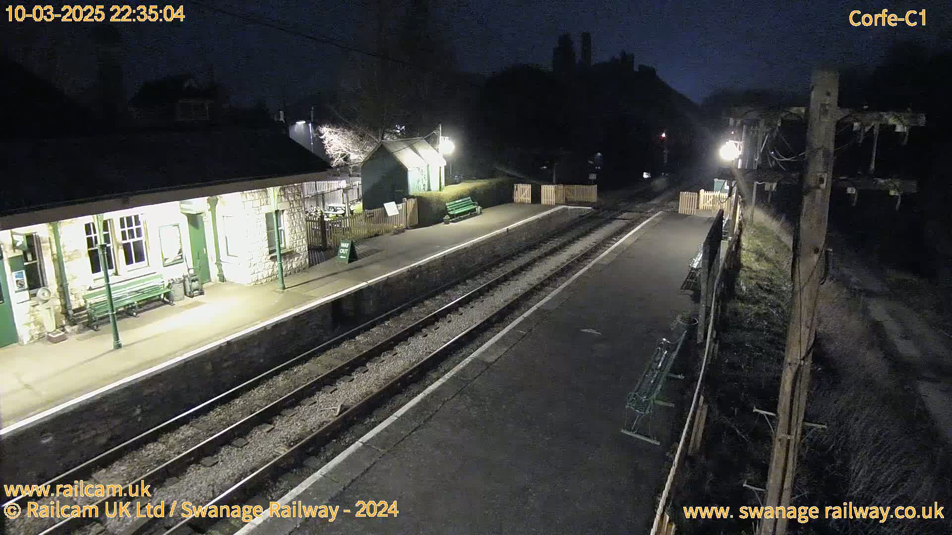 A nighttime view of a deserted train station platform with two sets of train tracks and a stone building.