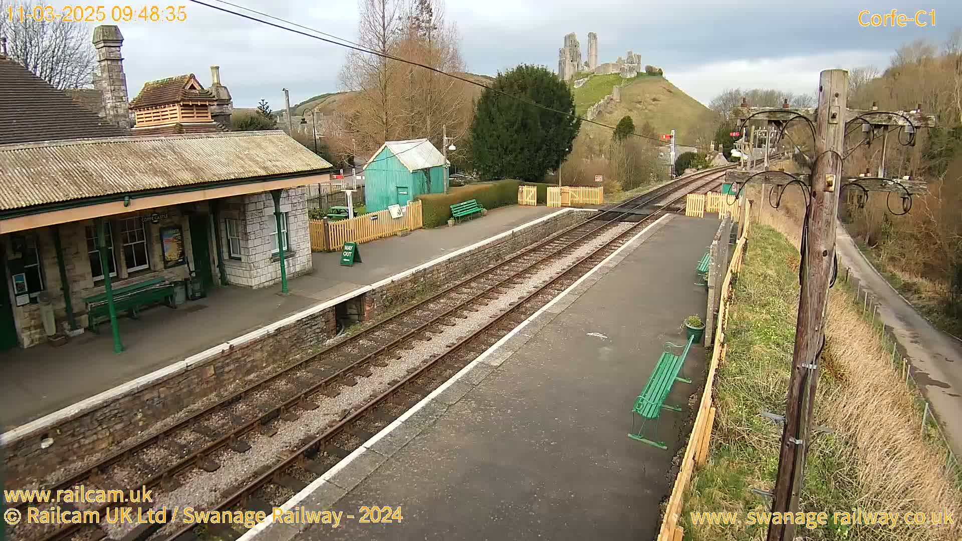 A train station platform with a stone building and benches, overlooking a hill with a castle ruin under an overcast sky.