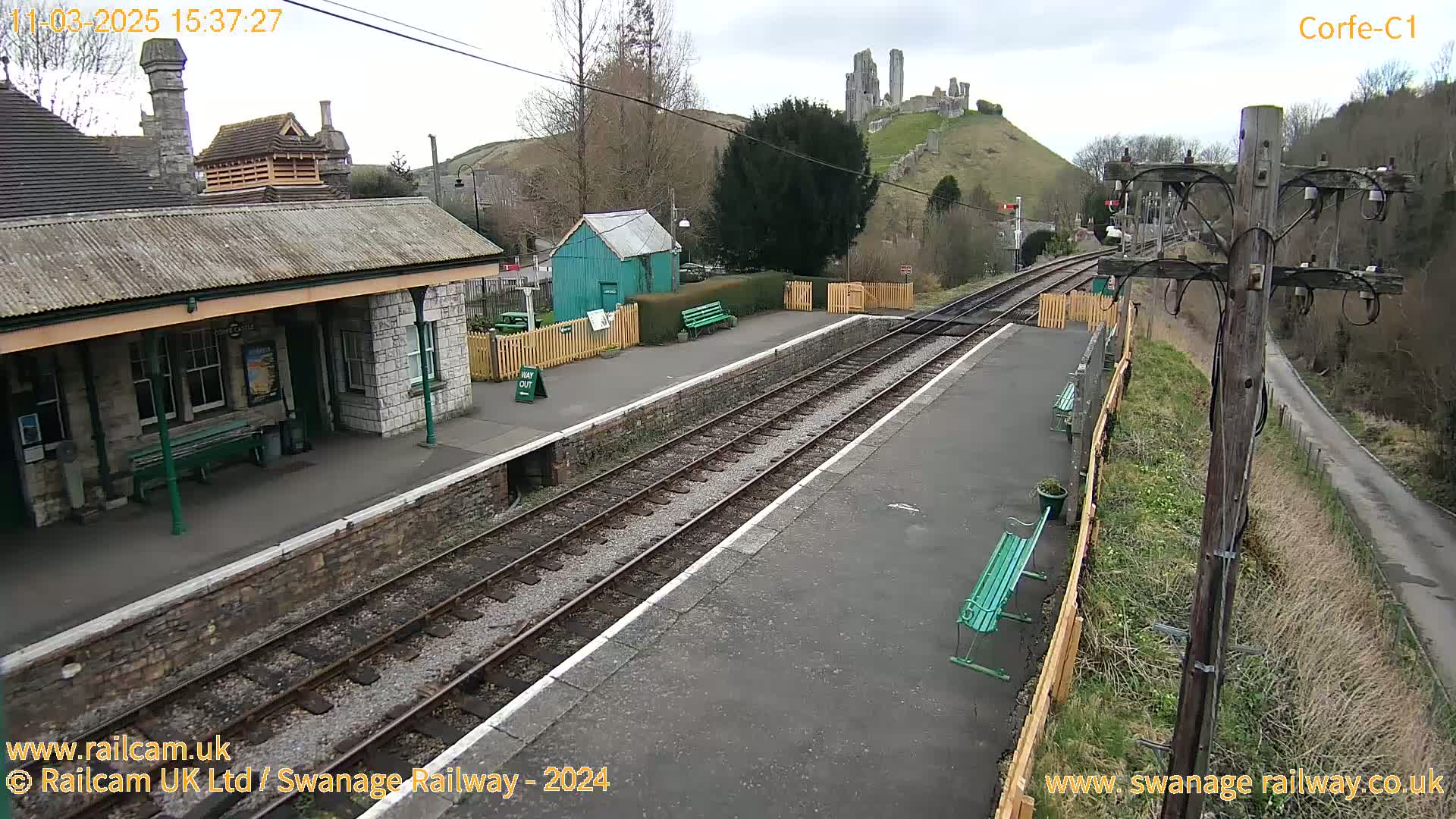 A train station with a stone building and platform, train tracks, and benches, overlooks a hill with castle ruins under an overcast sky.