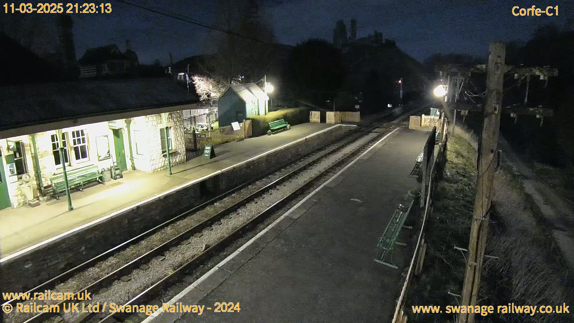 A nighttime view of a deserted train station platform with two sets of train tracks, illuminated by lights, and a stone building.