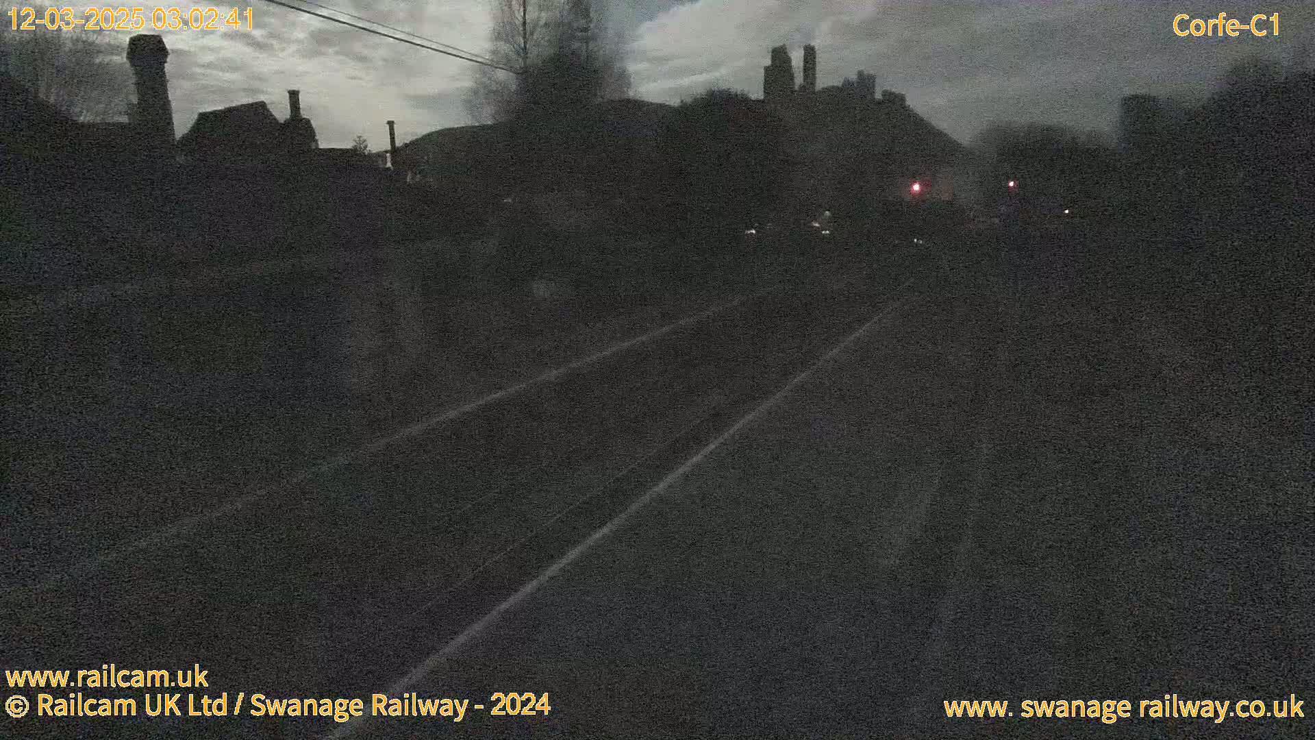 A dark, overcast view of a railway track at night, with buildings and trees silhouetted against a cloudy sky in the distance.