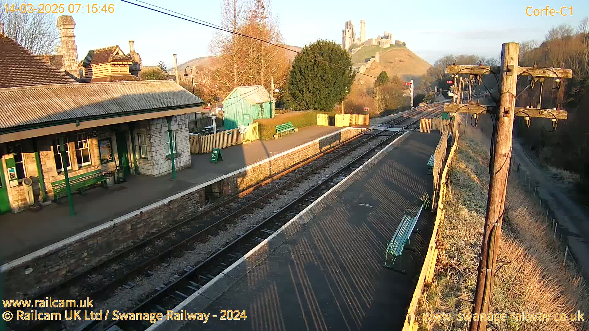 A train station platform with a stone building and a distant castle is visible under a sunny sky.