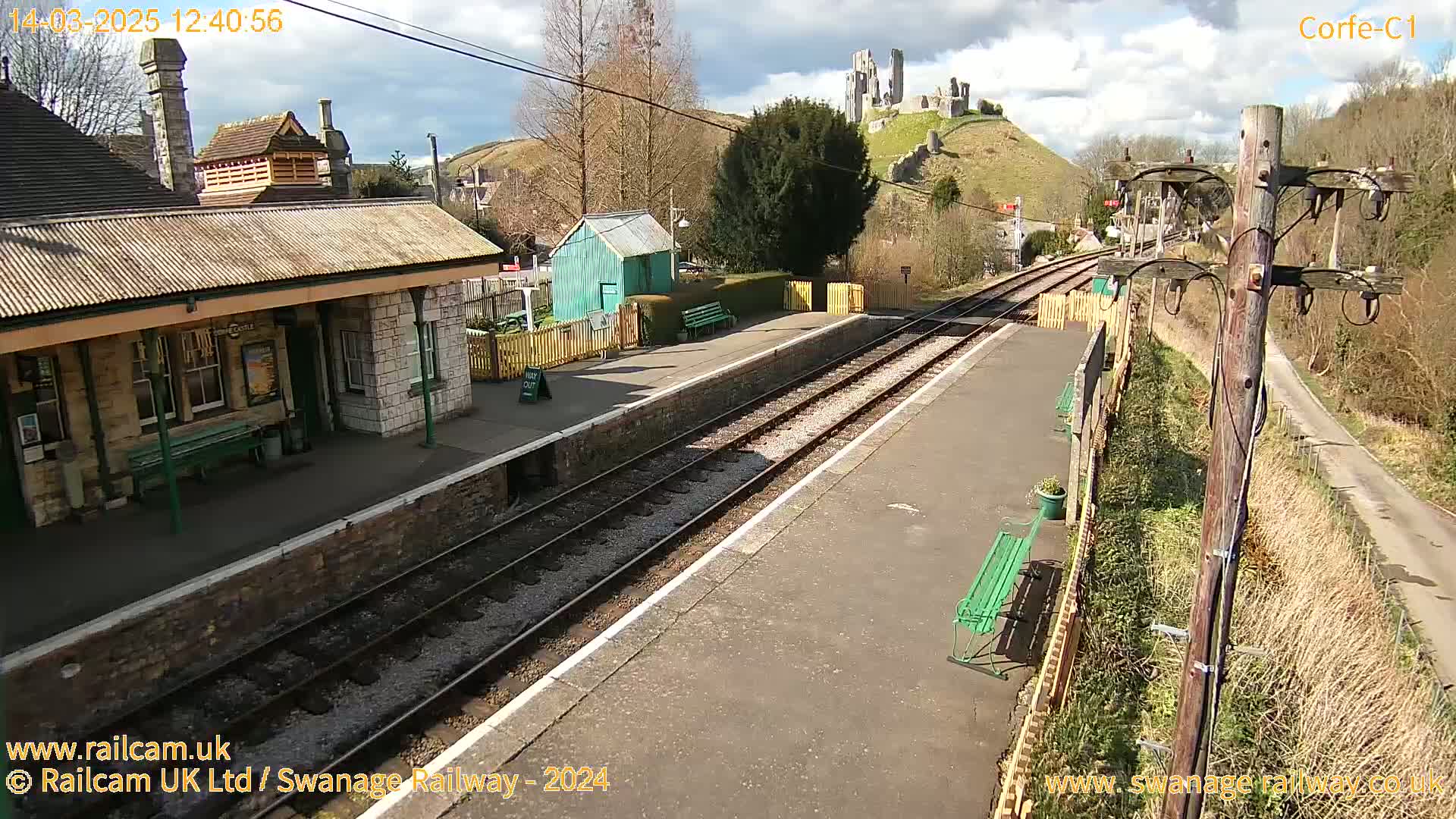 A train station platform with a stone building and a castle ruin visible on a hill in the background under a partly cloudy sky.