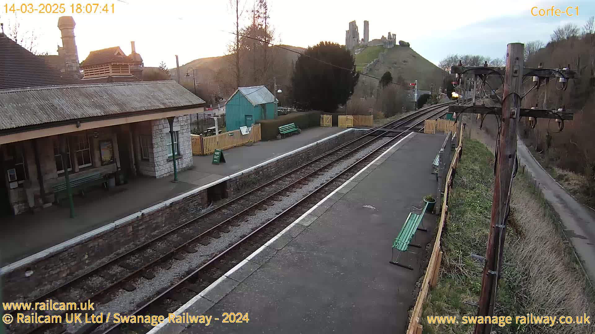 A train station with a stone building and benches, two train tracks, and a hill with castle ruins in the background under a partly cloudy sky.