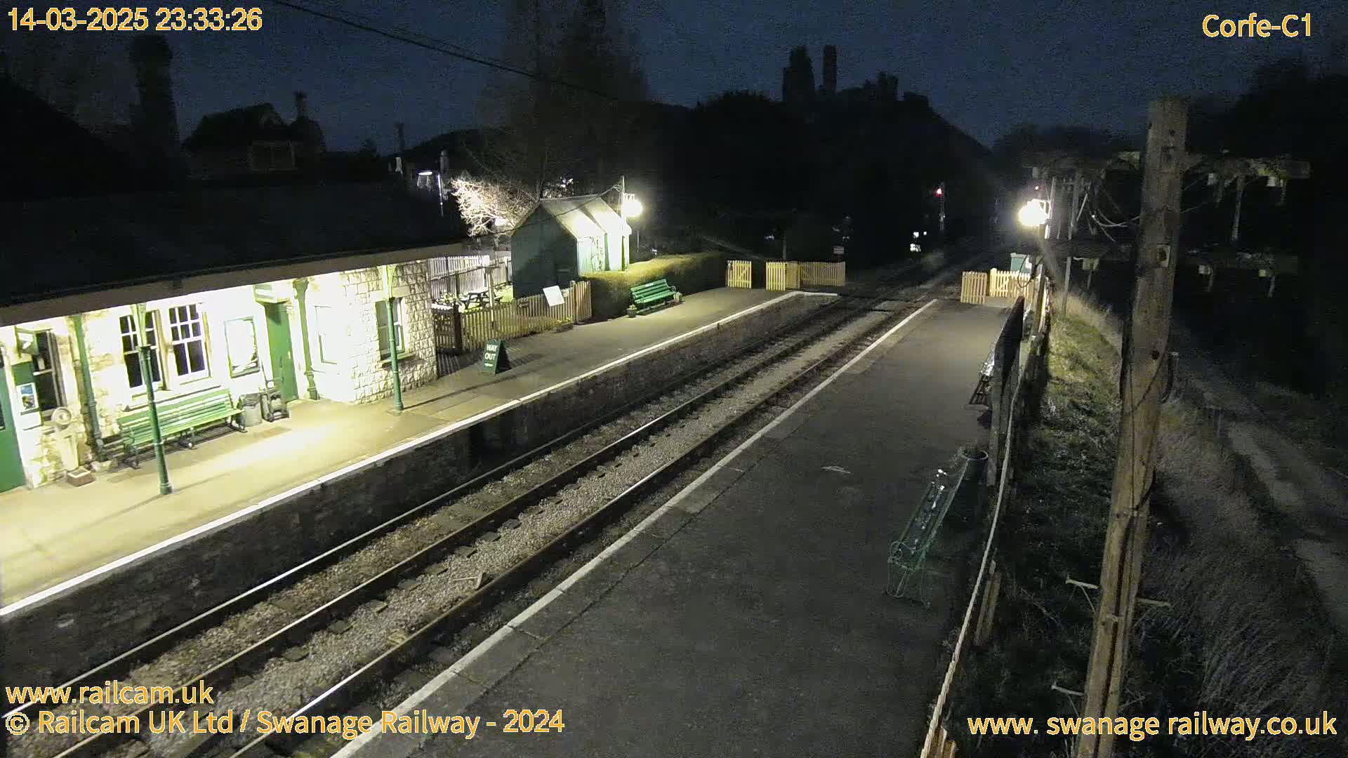 A nighttime view of a deserted train station platform with two sets of train tracks, illuminated by several lights, under a dark sky.