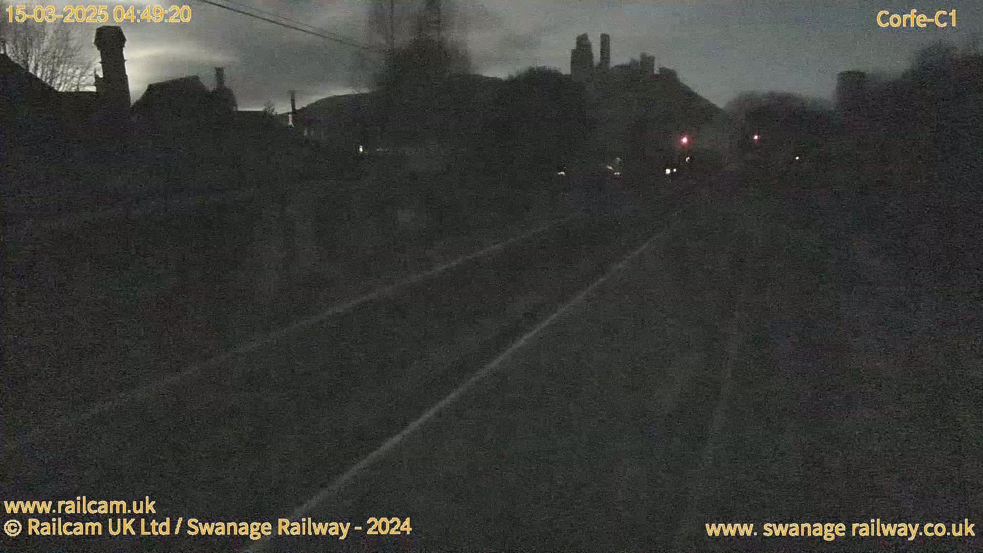 A dark, nighttime view of a railway track, with silhouetted buildings and trees in the background under a cloudy sky.
