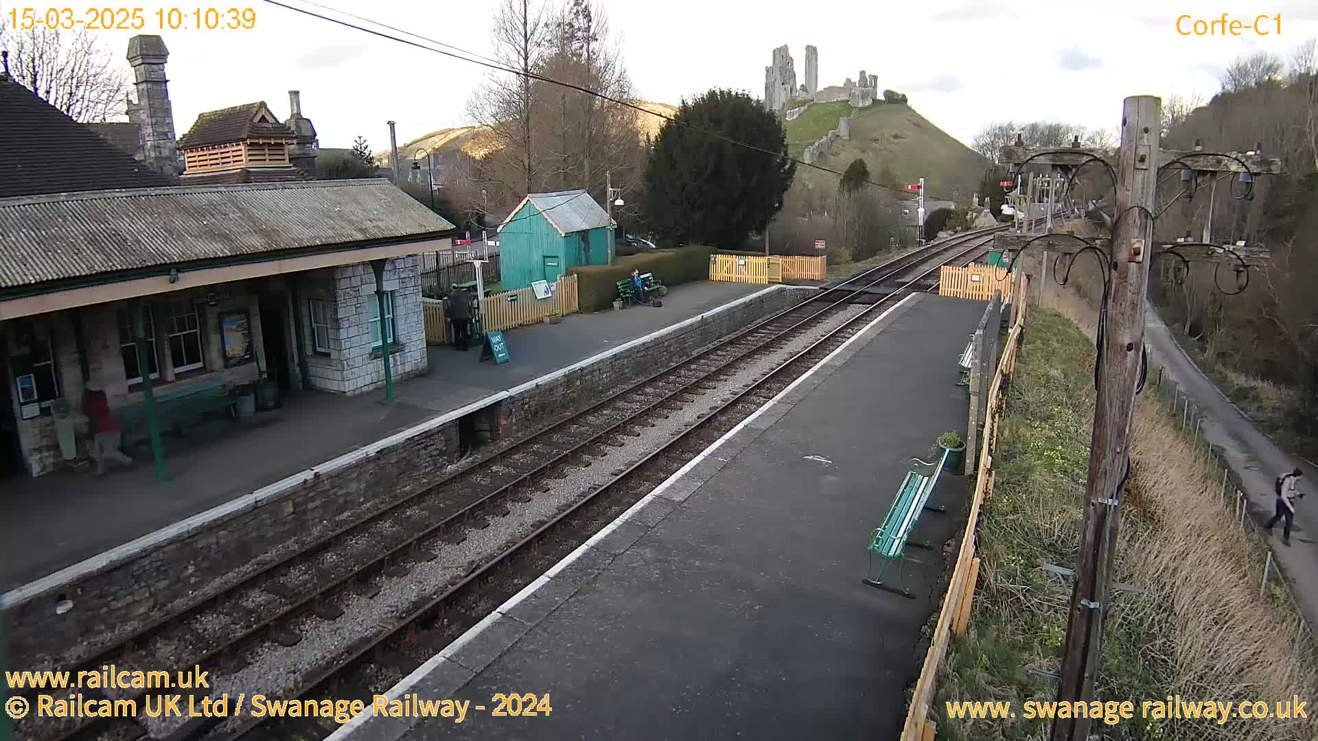 A train station with a stone building, a small green shed, and train tracks, all under a partly cloudy sky, with a hilltop castle visible in the background.