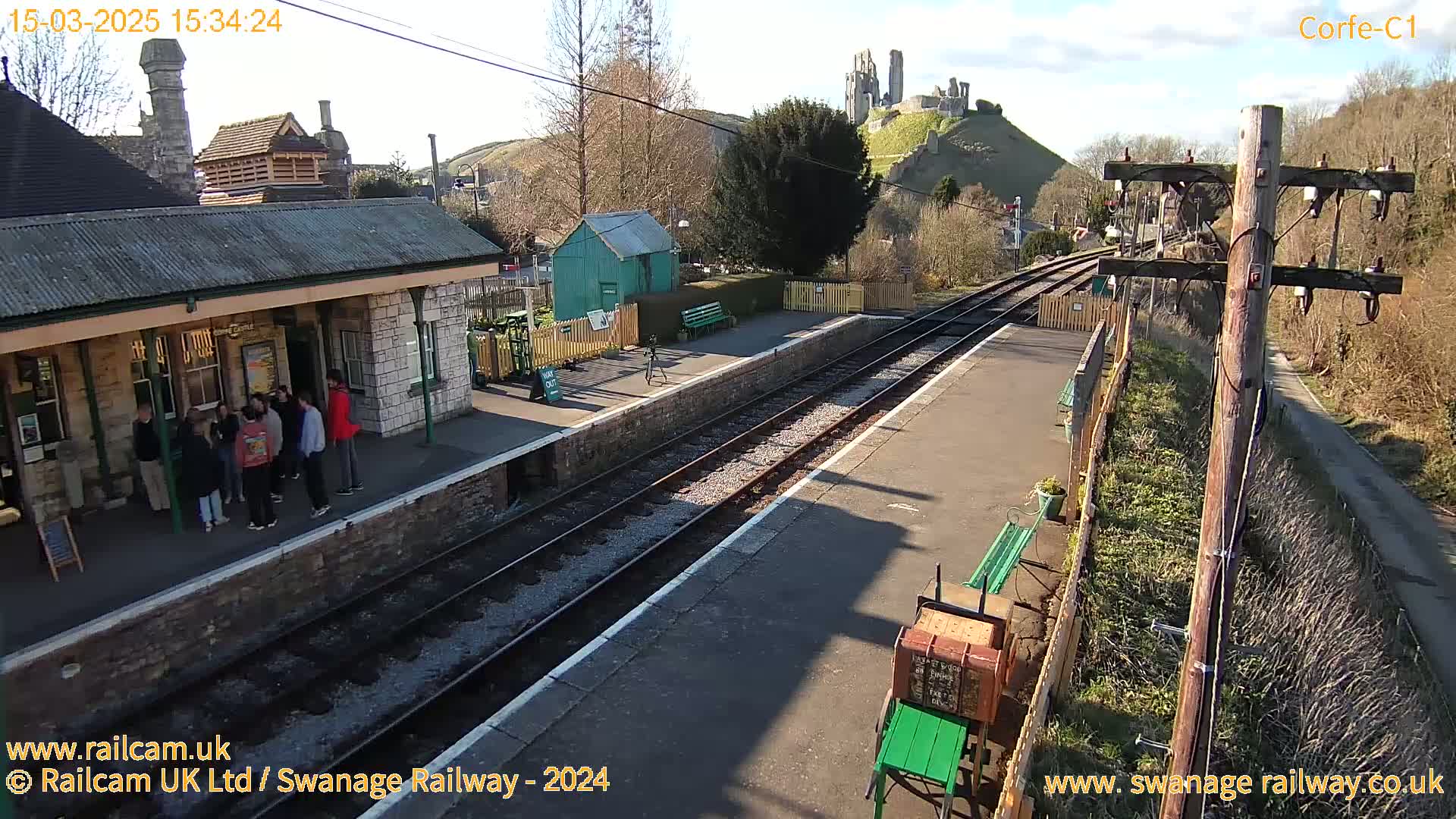 A train station platform on a sunny day, with a group of people standing near a stone building and a hilltop castle in the background.