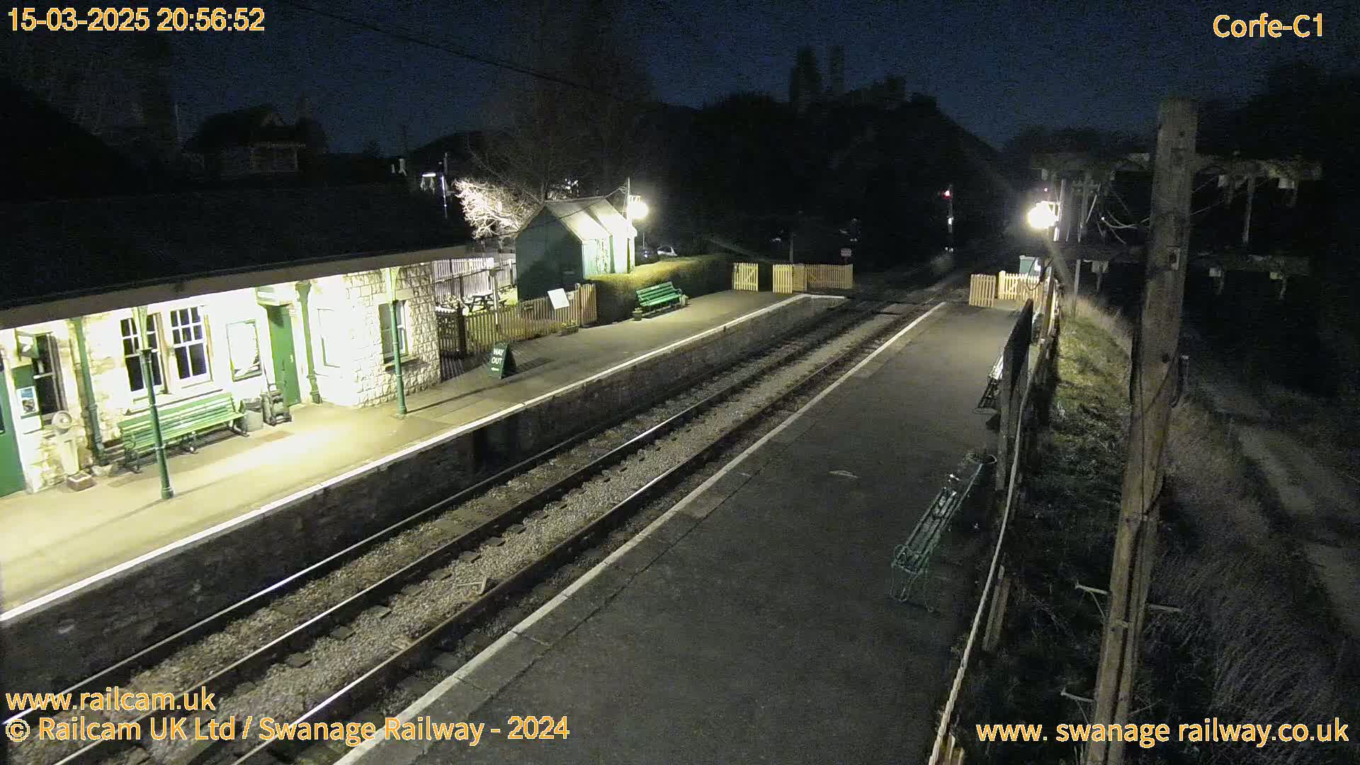 A deserted train station platform at night, illuminated by lights, with two sets of train tracks extending into the darkness.