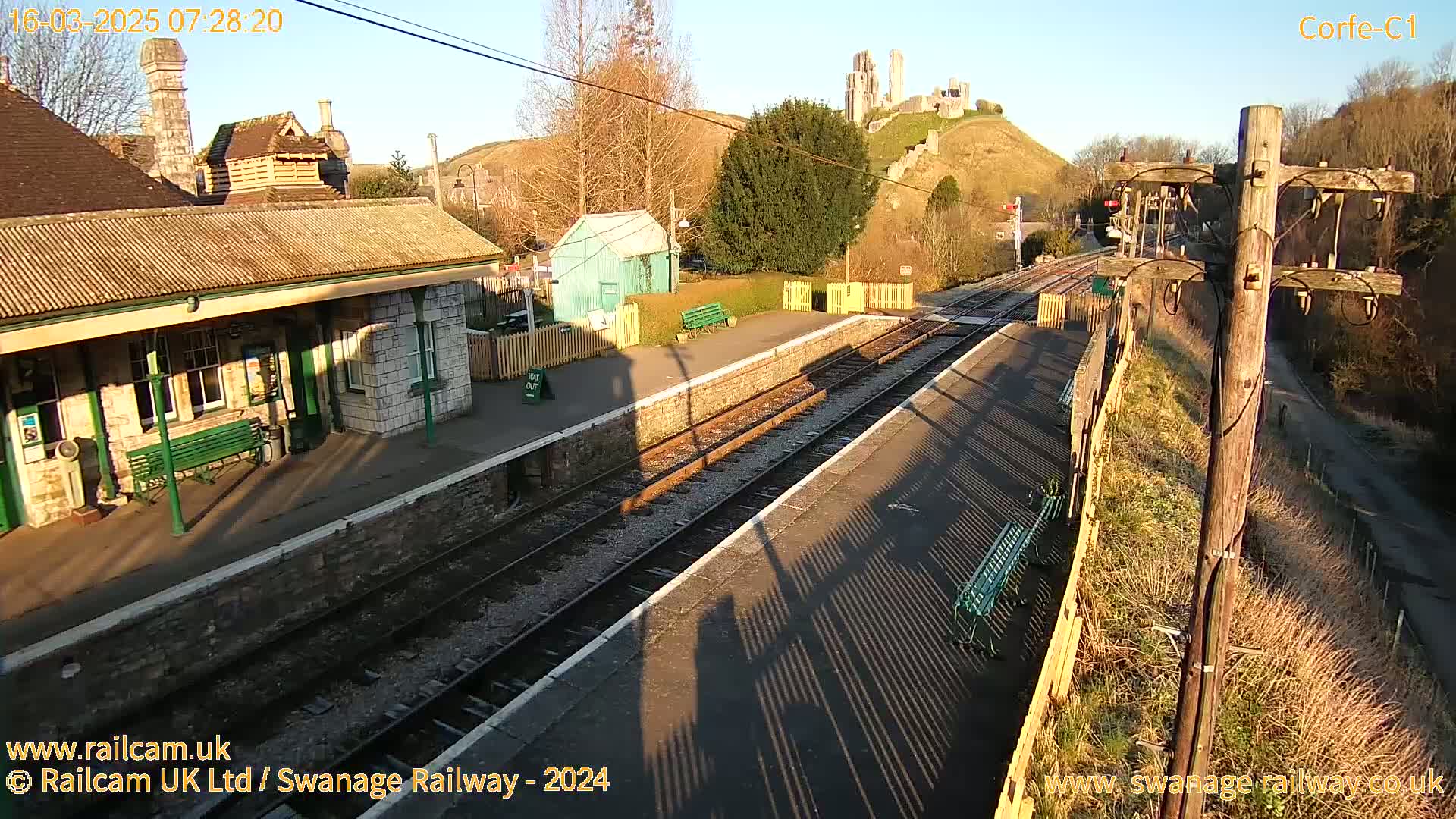 A train station platform with a stone building, railway tracks, and a castle visible in the distance on a sunny morning.