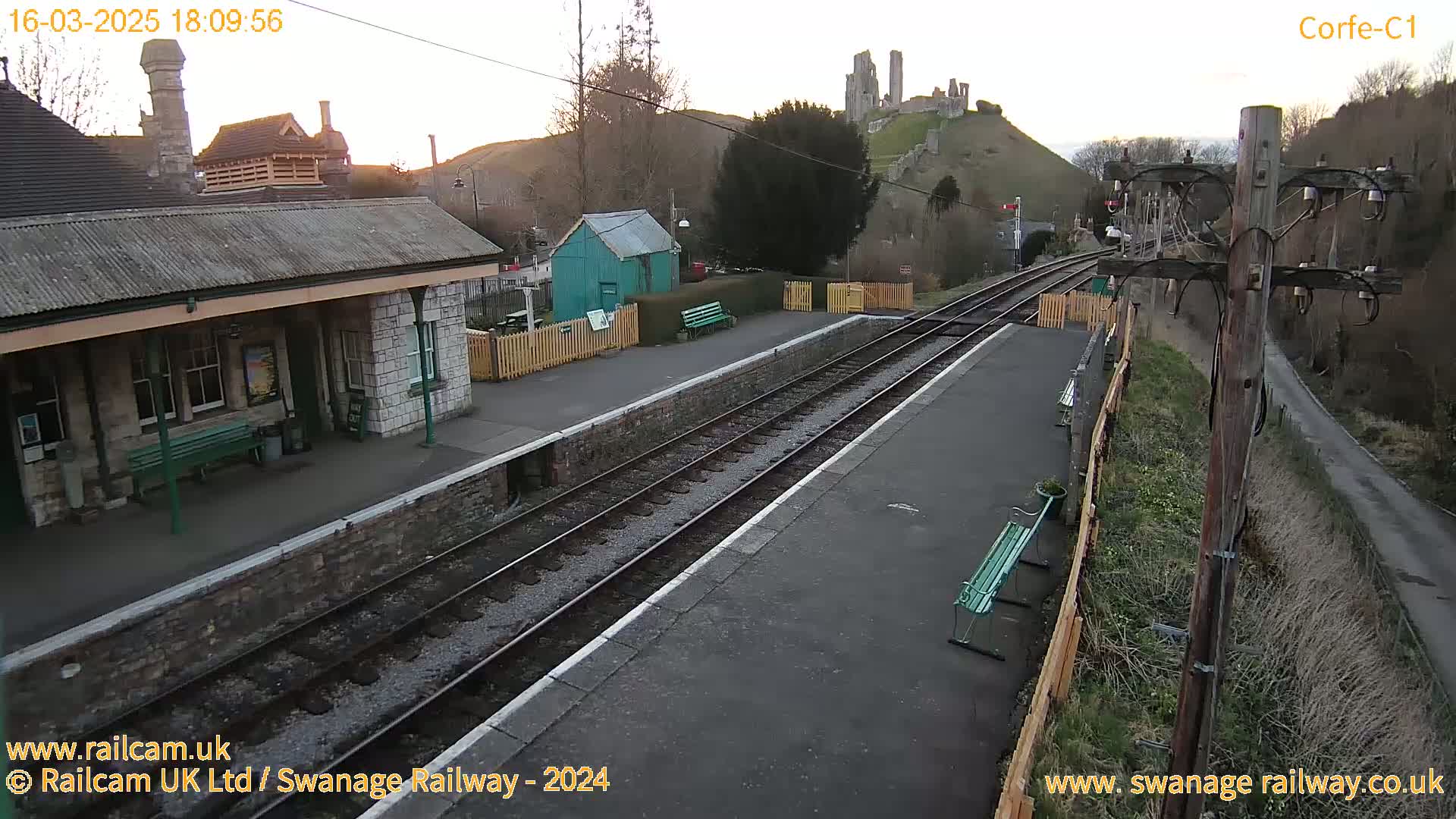 A train station platform with a stone building, a small green shed, and a hilltop castle ruin in the background under a twilight sky.