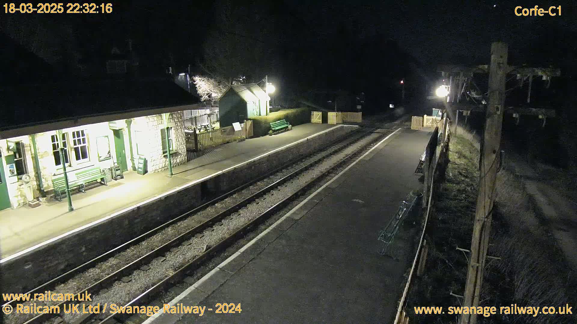 A nighttime view of a deserted train station platform with two sets of tracks, lit by lamps, under dark, clear skies.