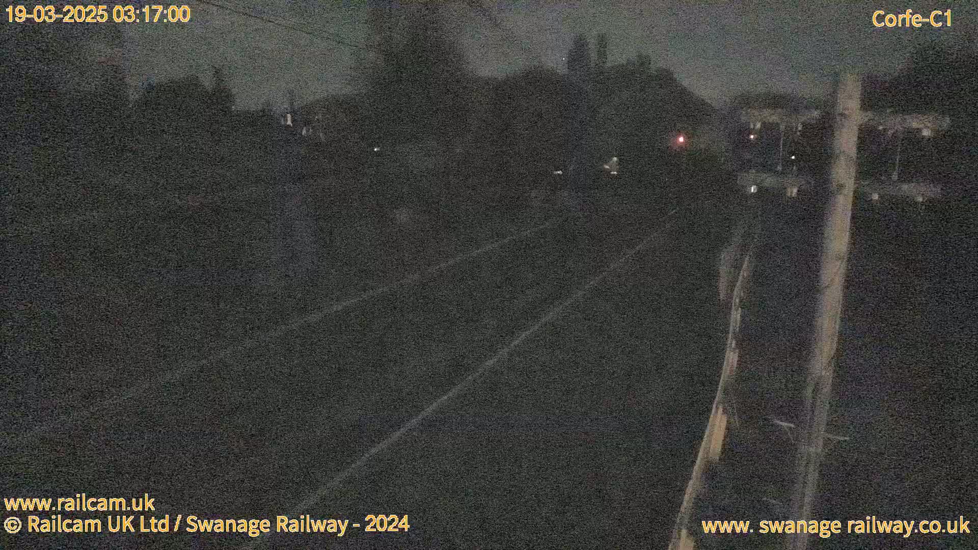 A nighttime view of a dark railway track with indistinct vegetation and structures in the background under a dark sky.