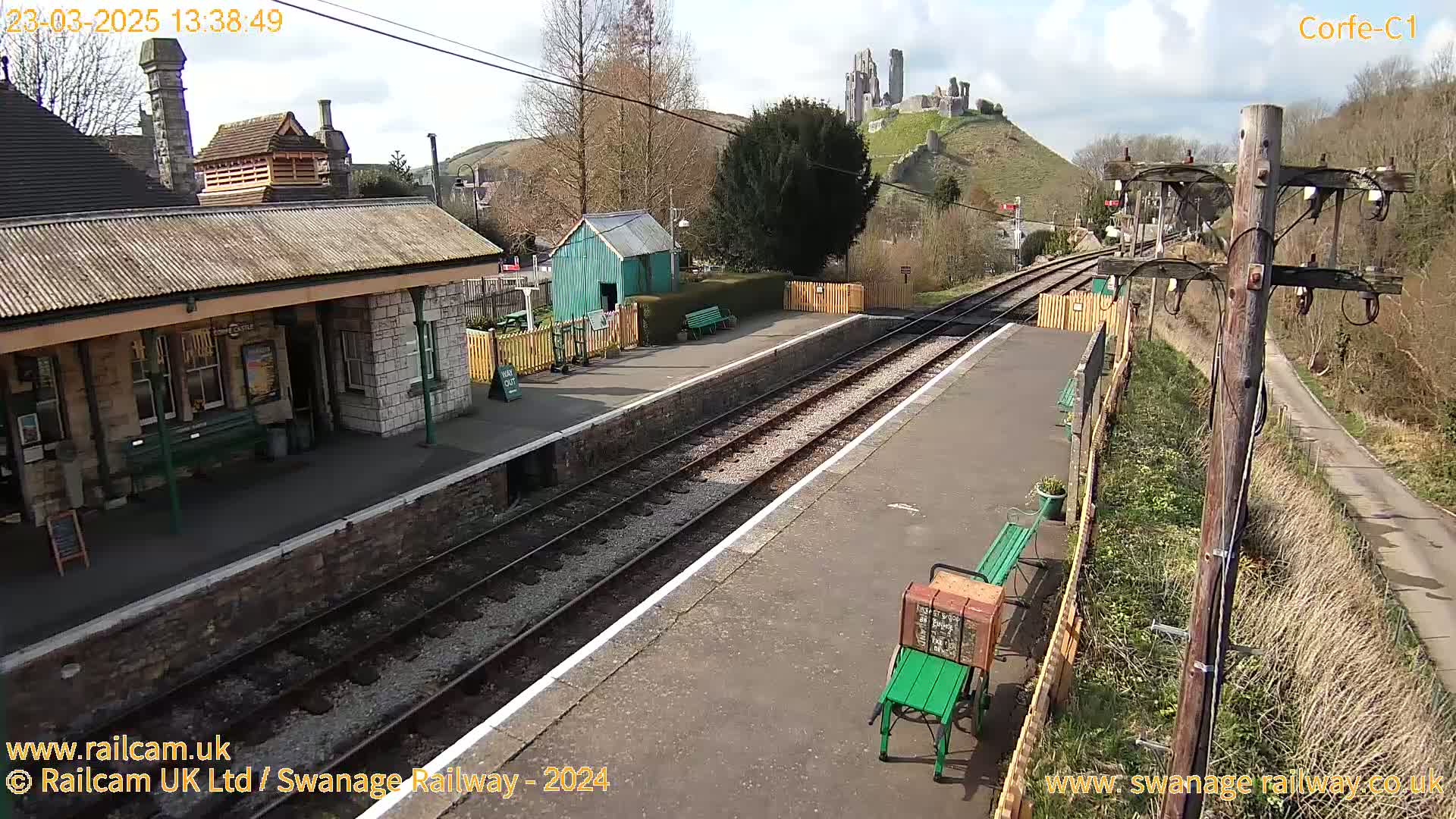 A train station platform with a stone building, a small green shed, and a castle visible on a hill in the background under a mostly sunny sky.