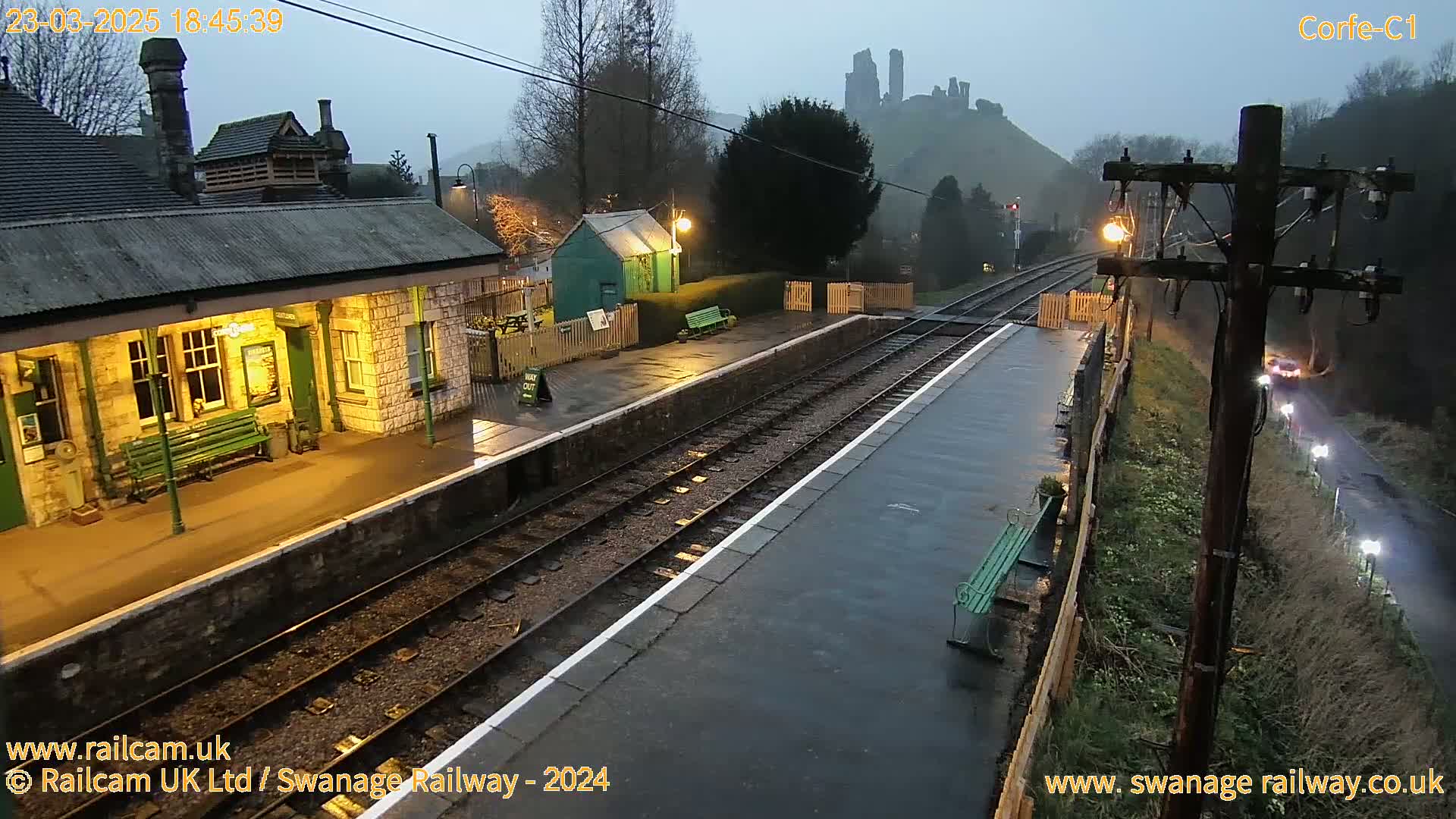 A wet, dimly lit train station platform at dusk, with a stone building and a distant castle visible on a hill.