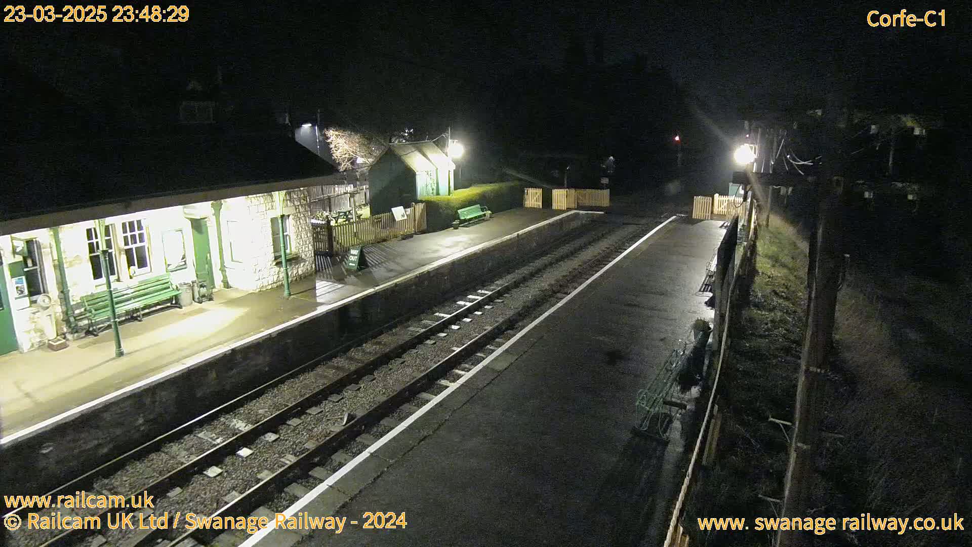 A nighttime view of a train station platform with two sets of tracks, a building, and lights illuminating the scene.