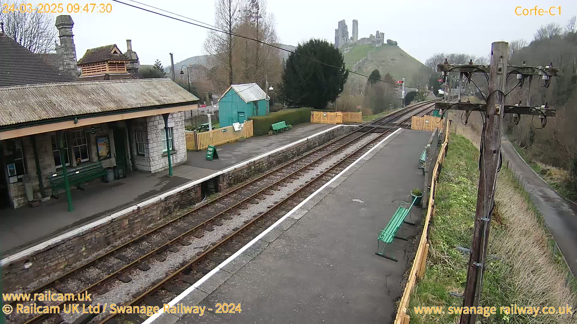 A train station with a stone building and a small green shed sits beside train tracks, with a castle visible on a hill in the background under an overcast sky.