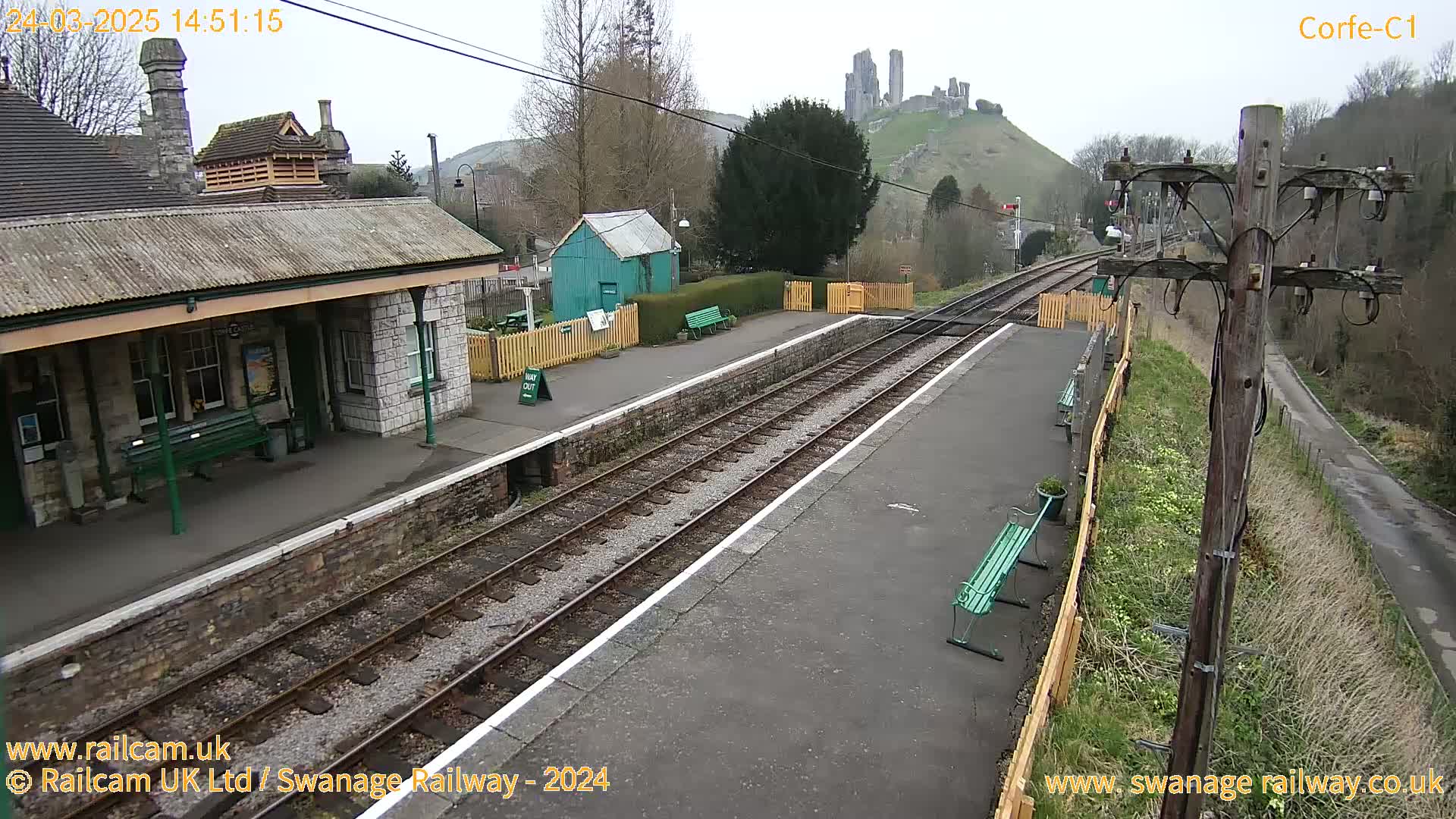 A train station platform with a stone building and a hilltop castle in the background on an overcast day.