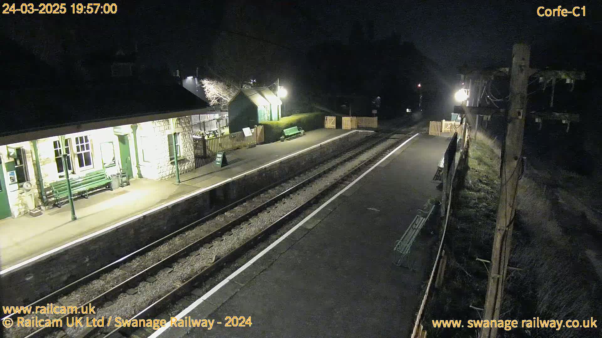 A deserted train station platform at night, illuminated by lights, with train tracks and a building in the background.