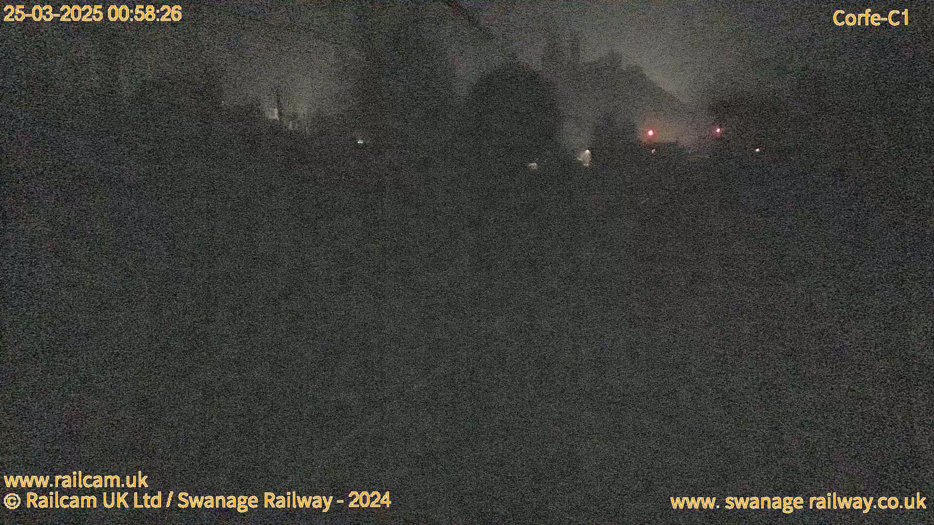 A nighttime, dark outdoor scene shows indistinct shapes in the distance, possibly buildings or trees, under a cloudy or hazy sky.